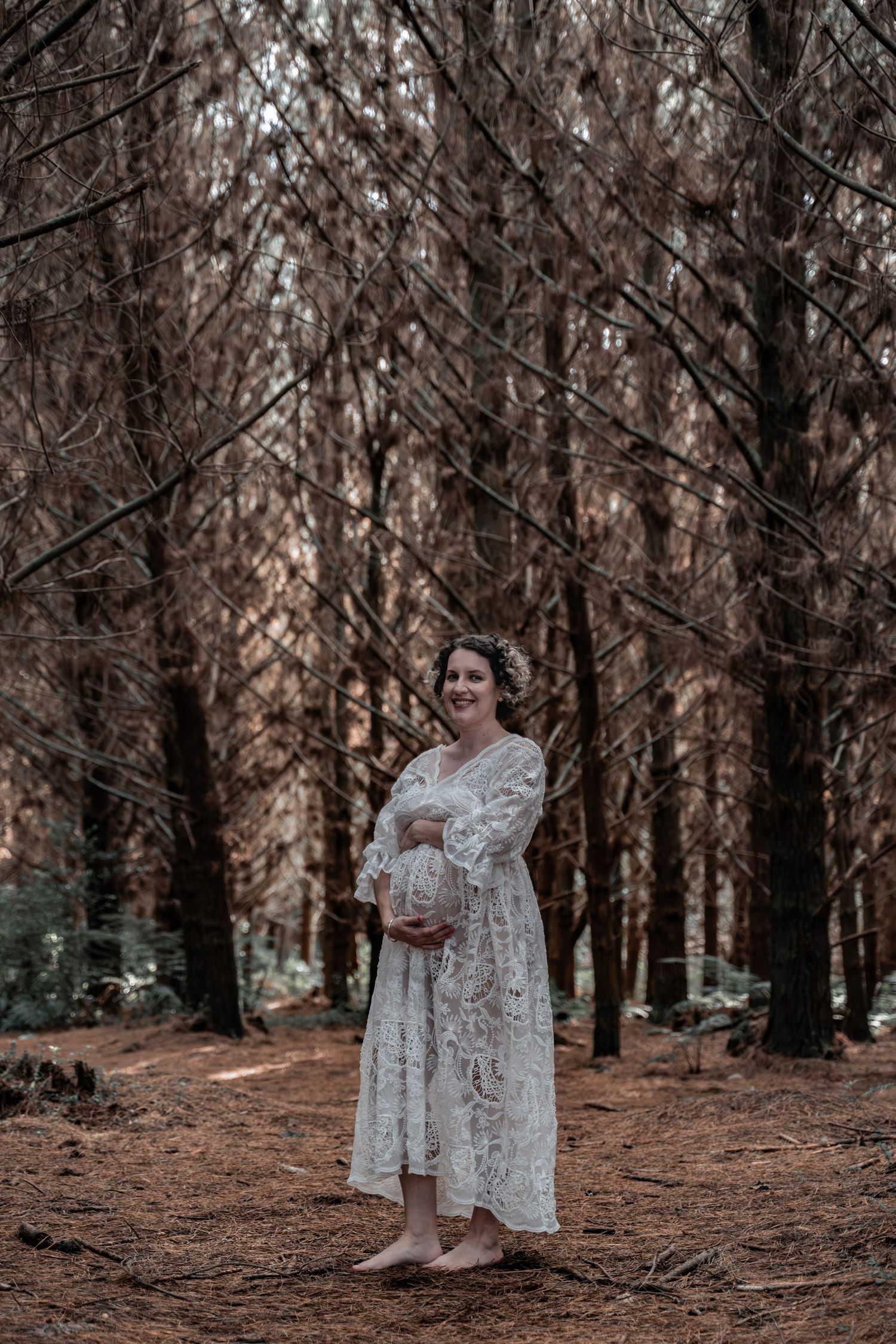 Pregnant woman in white lace dress holding baby bump, smiling in a forest with tall bare trees.