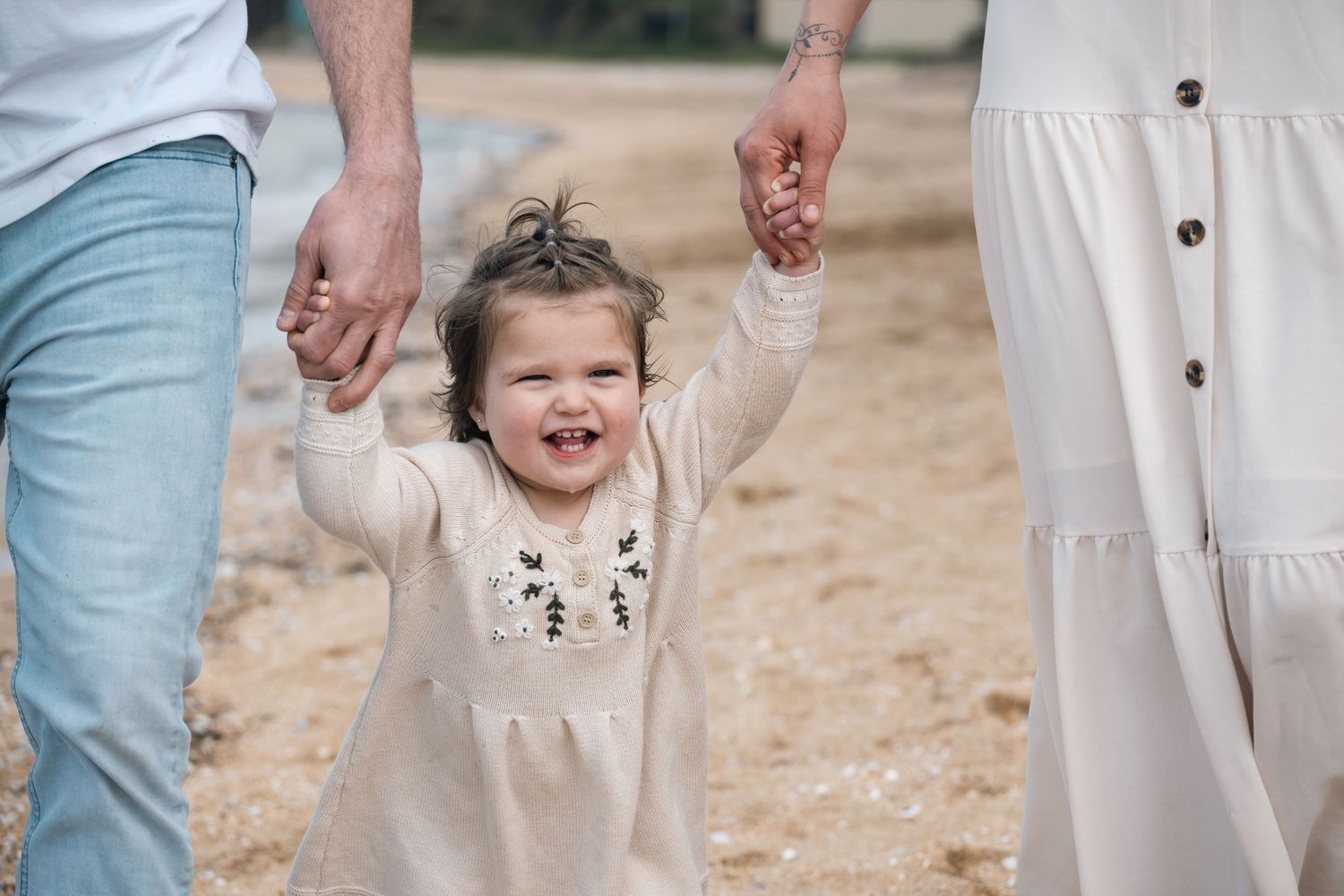 Smiling toddler girl in cream dress joyfully holds hands with parents outdoors on a sunny day.