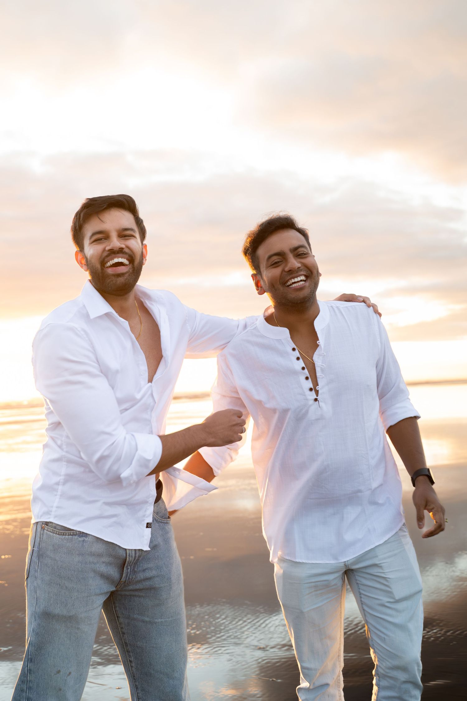 Two smiling men in white shirts walk together on a beach at sunset, one with arm around the other.