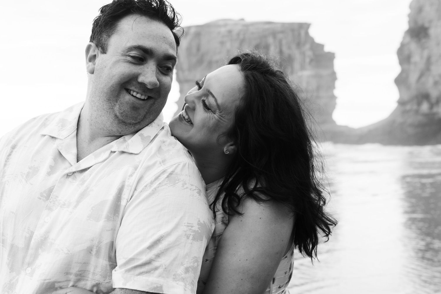 Black and white photo of a smiling couple embracing outdoors near water with a rocky cliff formation behind them.