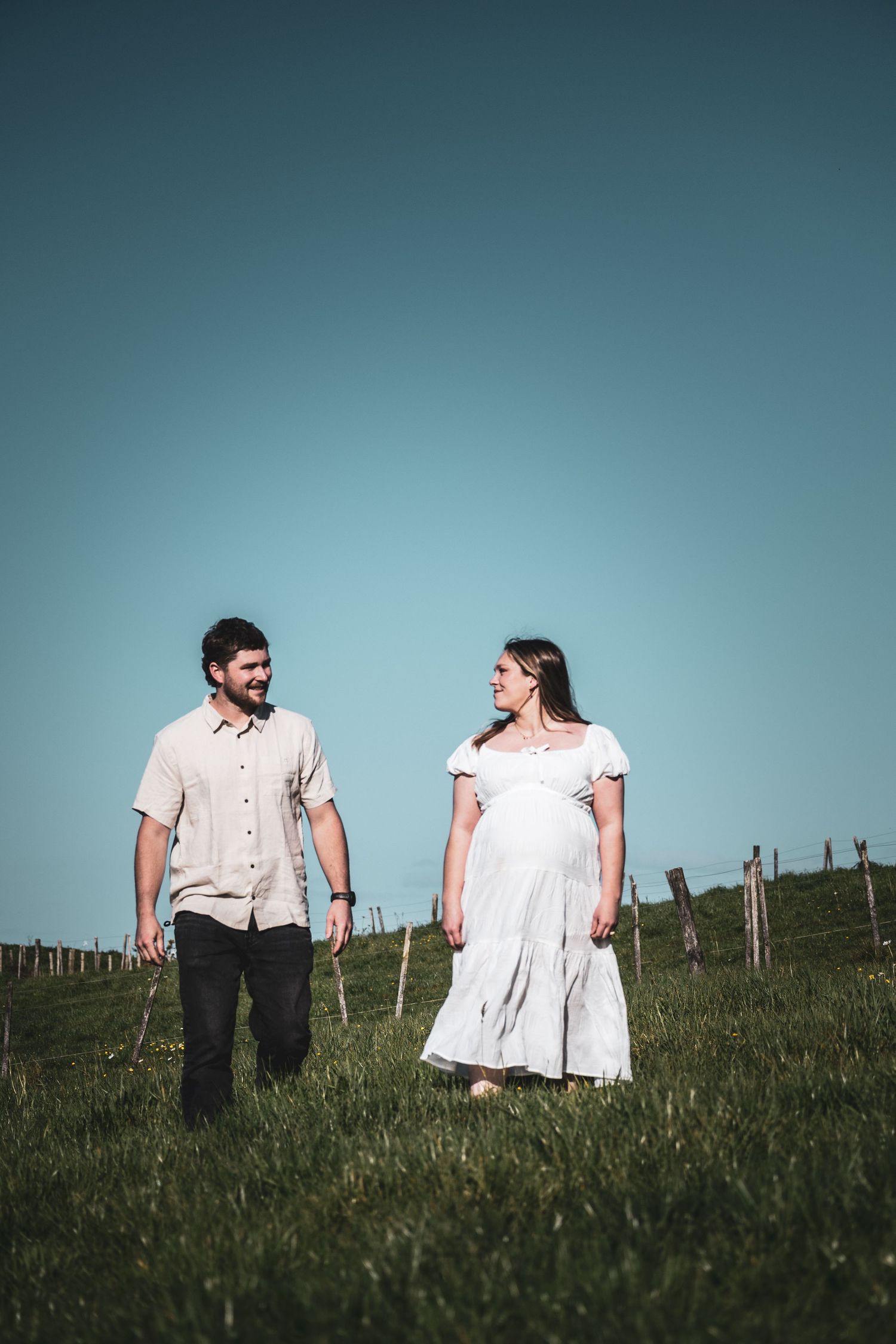 Pregnant woman in white dress and man in beige shirt walking together on green hillside under clear blue sky.