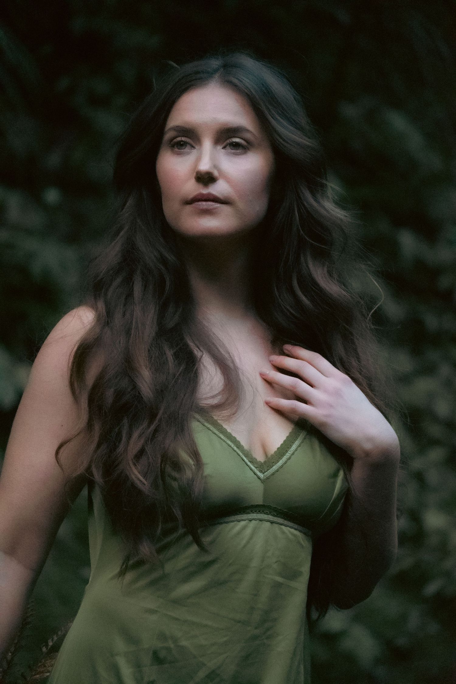 A woman with long dark wavy hair wearing a green dress poses outdoors against a lush dark green foliage background.