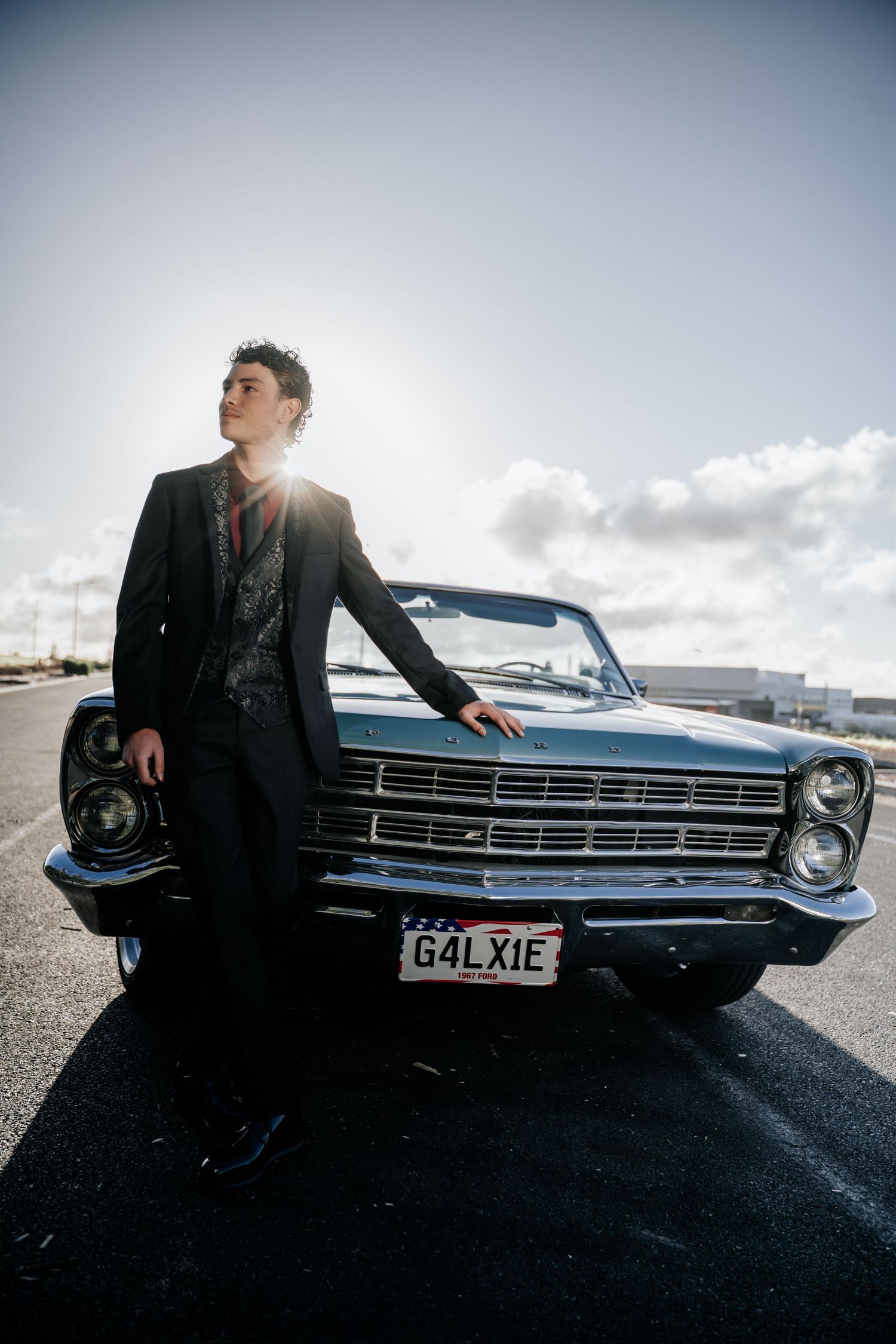 Young man in black suit leaning on vintage blue Ford Galaxie convertible in sunny parking lot.