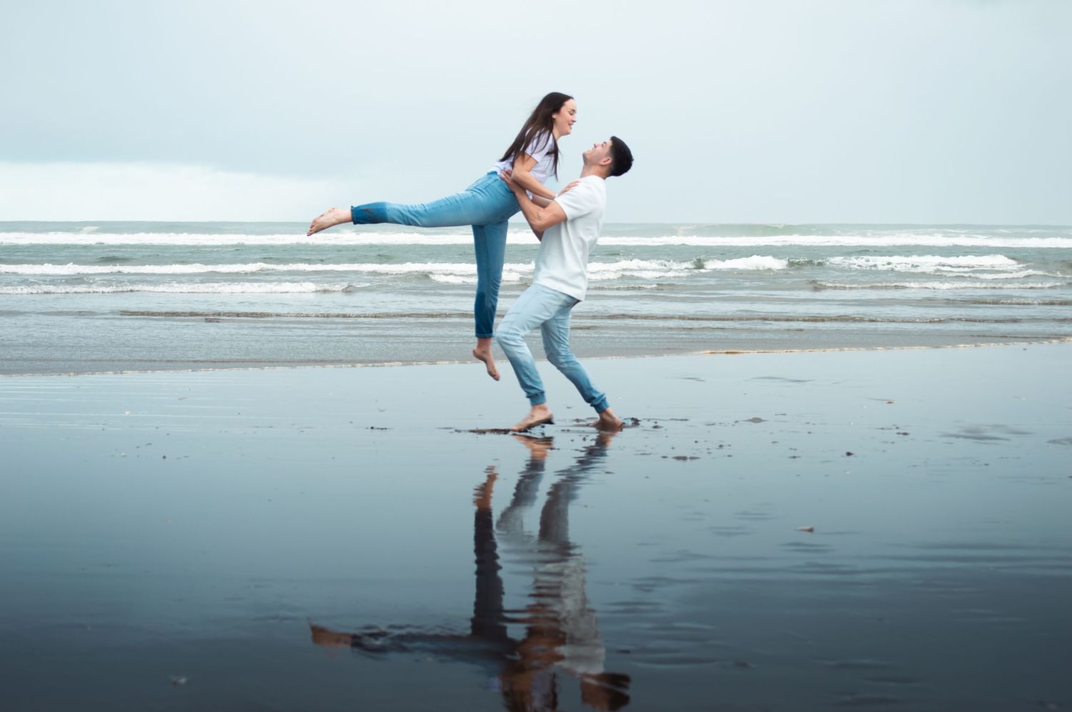 Couple dancing romantically on a reflective wet beach, man lifting woman in jeans against ocean waves.