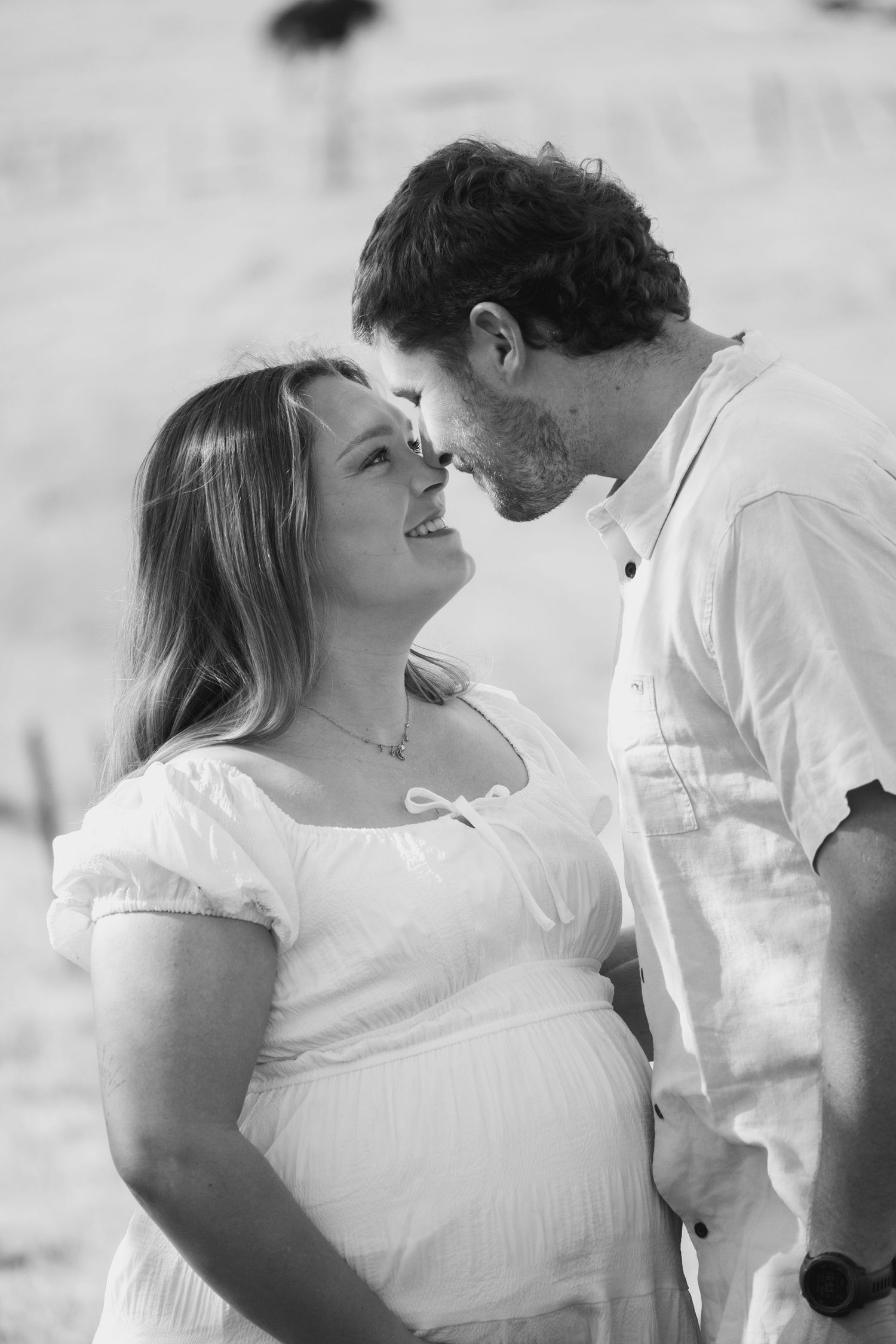 Black and white photo of a smiling pregnant woman in white dress with her partner leaning in close outdoors.
