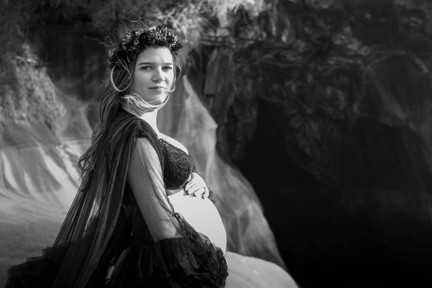 Black and white maternity photo of elegant pregnant woman in dark gown near rocky waterfall backdrop.
