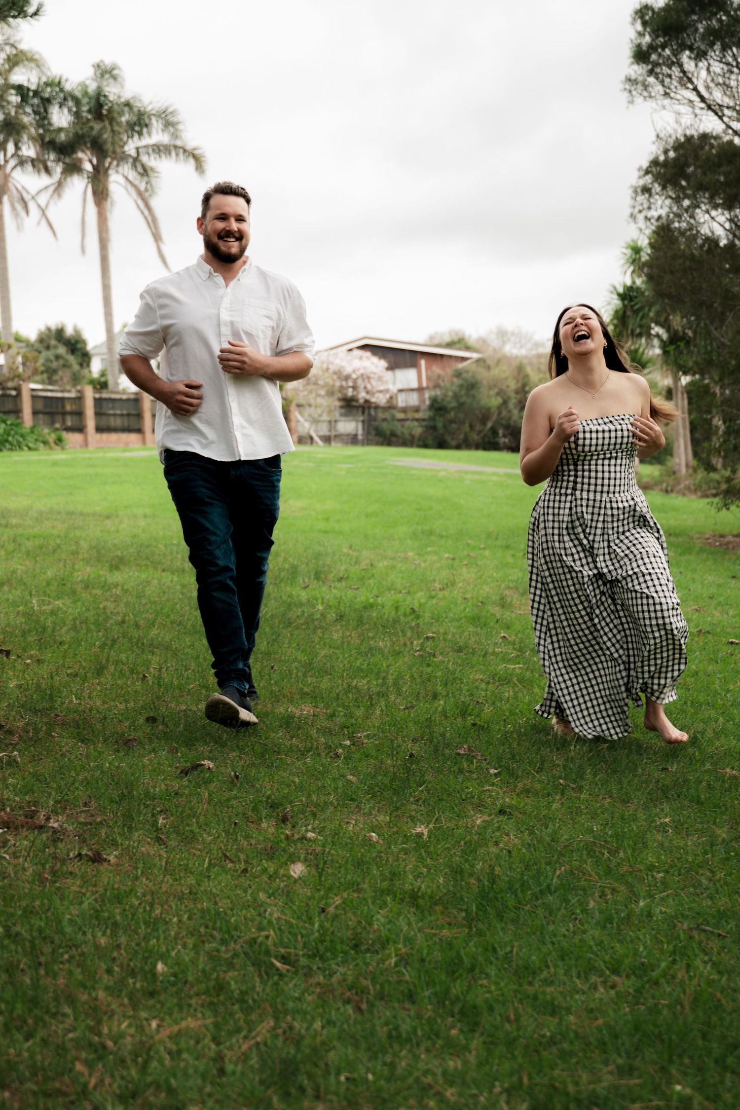 A laughing couple runs through a lush green park, woman in checkered dress and man in white shirt and jeans.