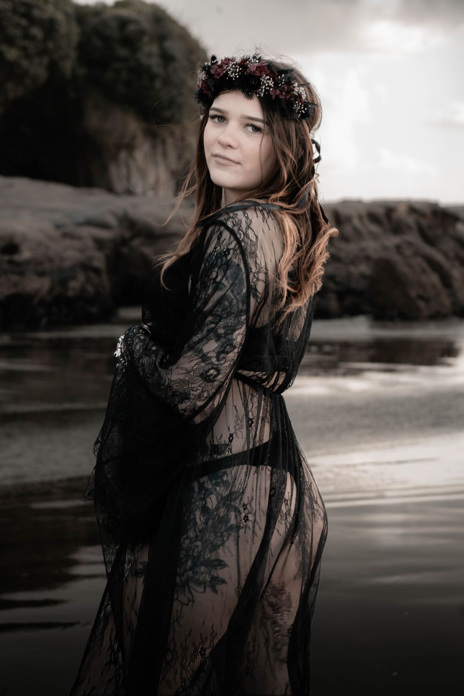Woman in black lace dress and floral crown poses gracefully by rocky shoreline at dusk.