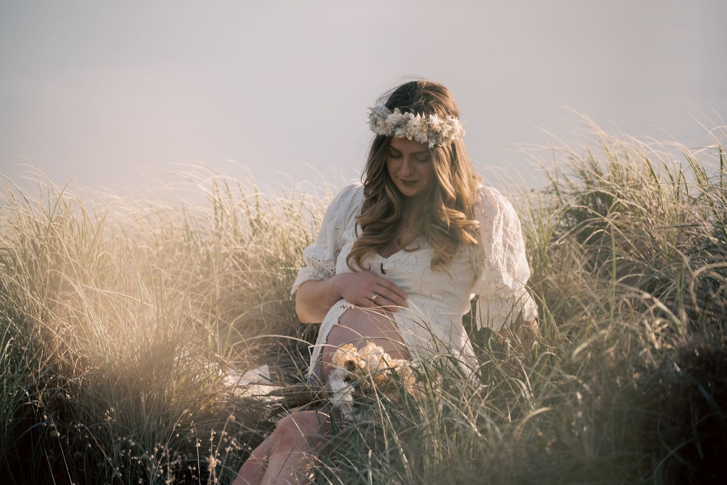 Pregnant woman in white dress and floral crown sitting in golden grass field during soft sunlight.