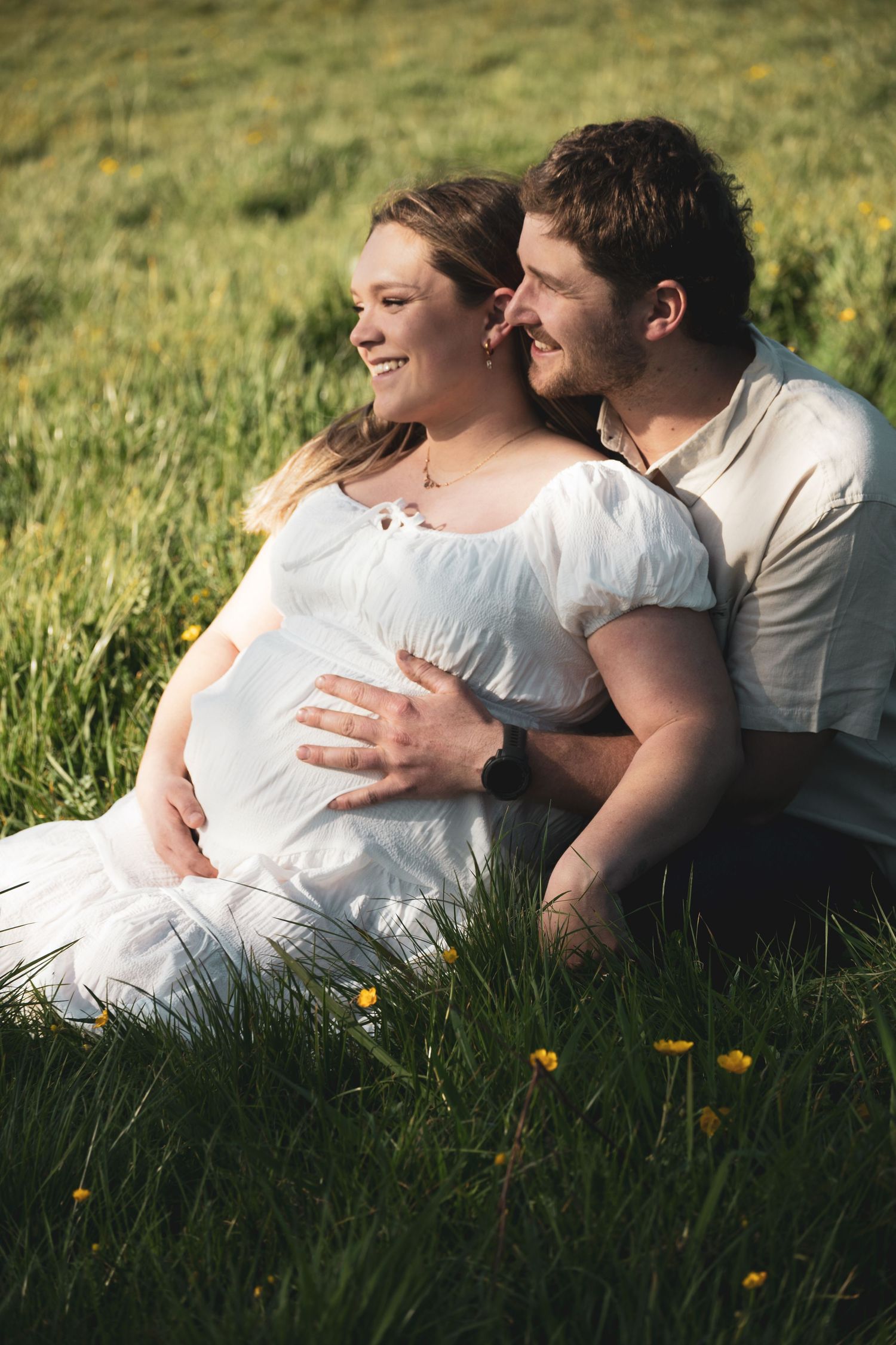 Pregnant couple sitting in green meadow with wildflowers, woman in white dress smiling as partner embraces her baby bump.