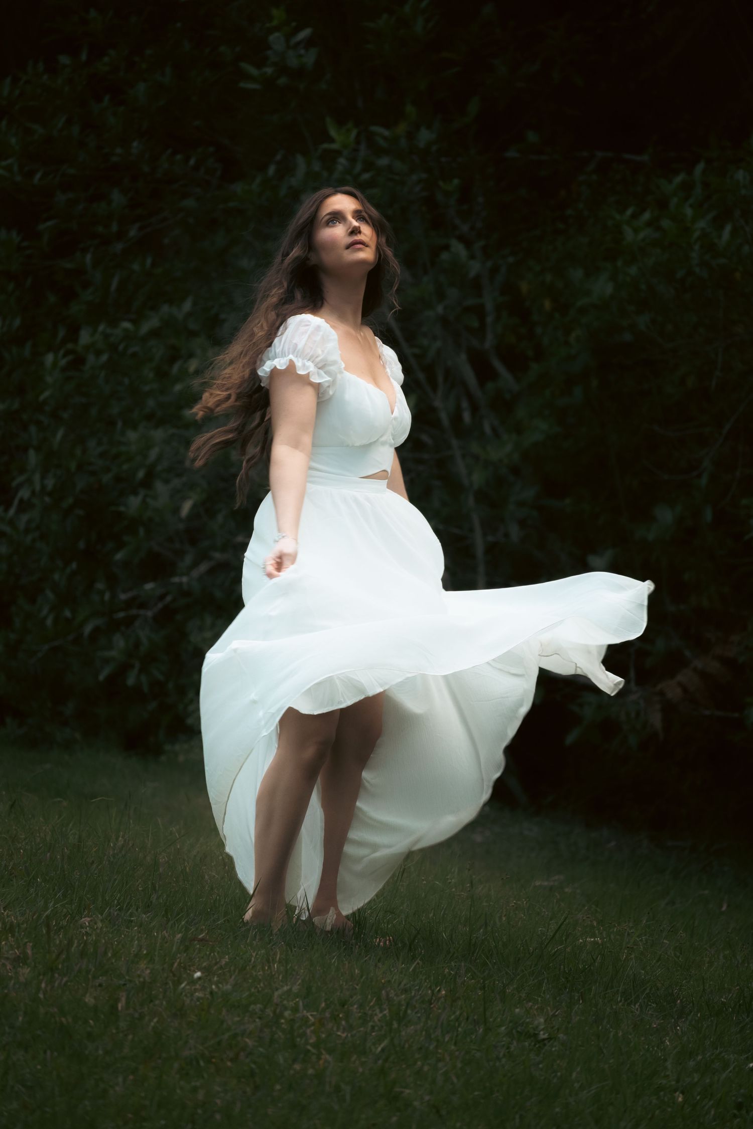 Woman in flowing white dress standing barefoot in dark garden, long hair blowing in wind, gazing upward.