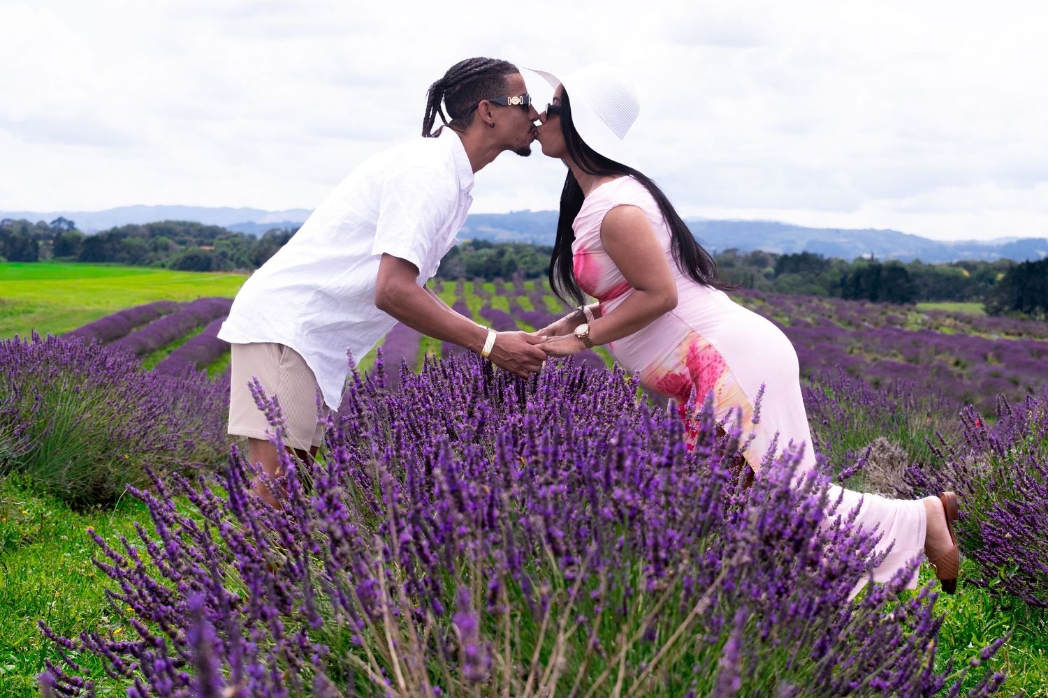 Couple sharing a romantic kiss in a vibrant purple lavender field, woman in pink dress, man in white outfit.