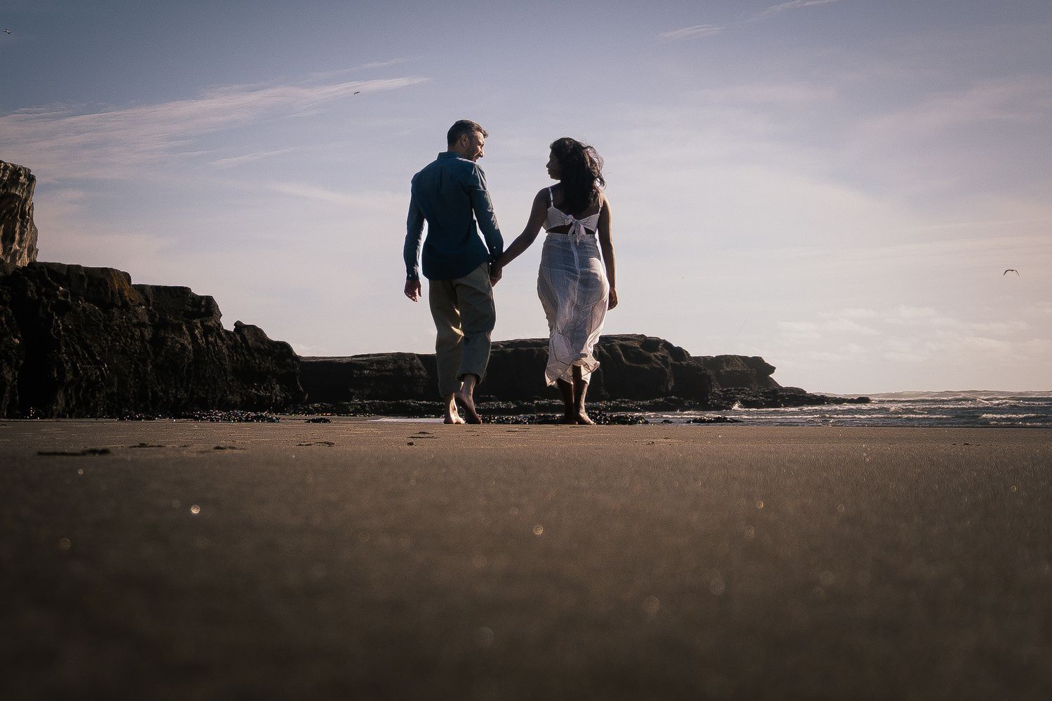 Couple holding hands walking barefoot on a sandy beach toward rocky cliffs at sunset.