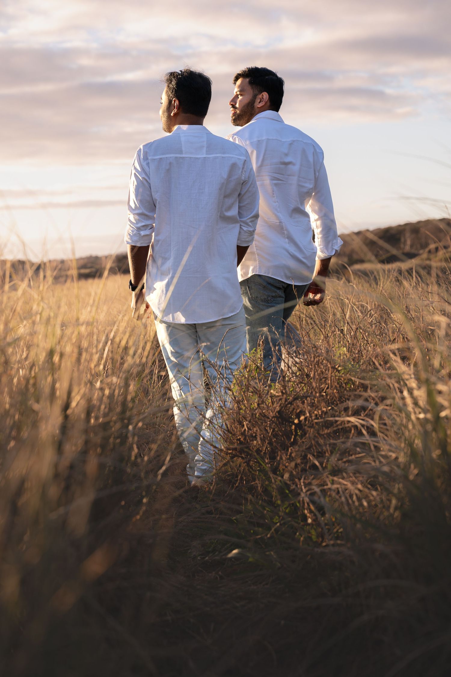 Two men in white shirts and jeans walk through golden field at sunset.
