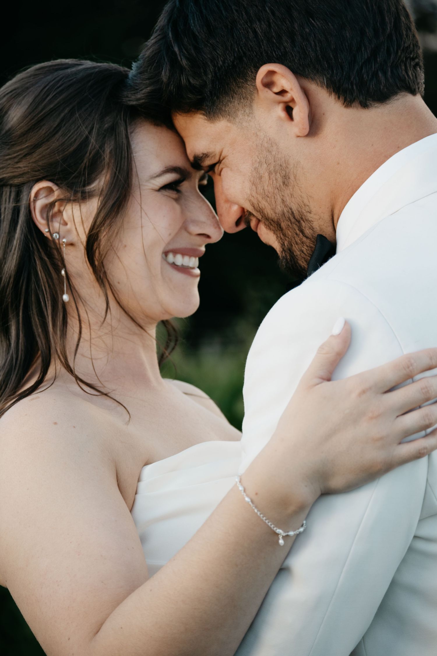 Smiling bride and groom share an intimate forehead touch moment, with the bride in a white dress wearing pearl earrings.
