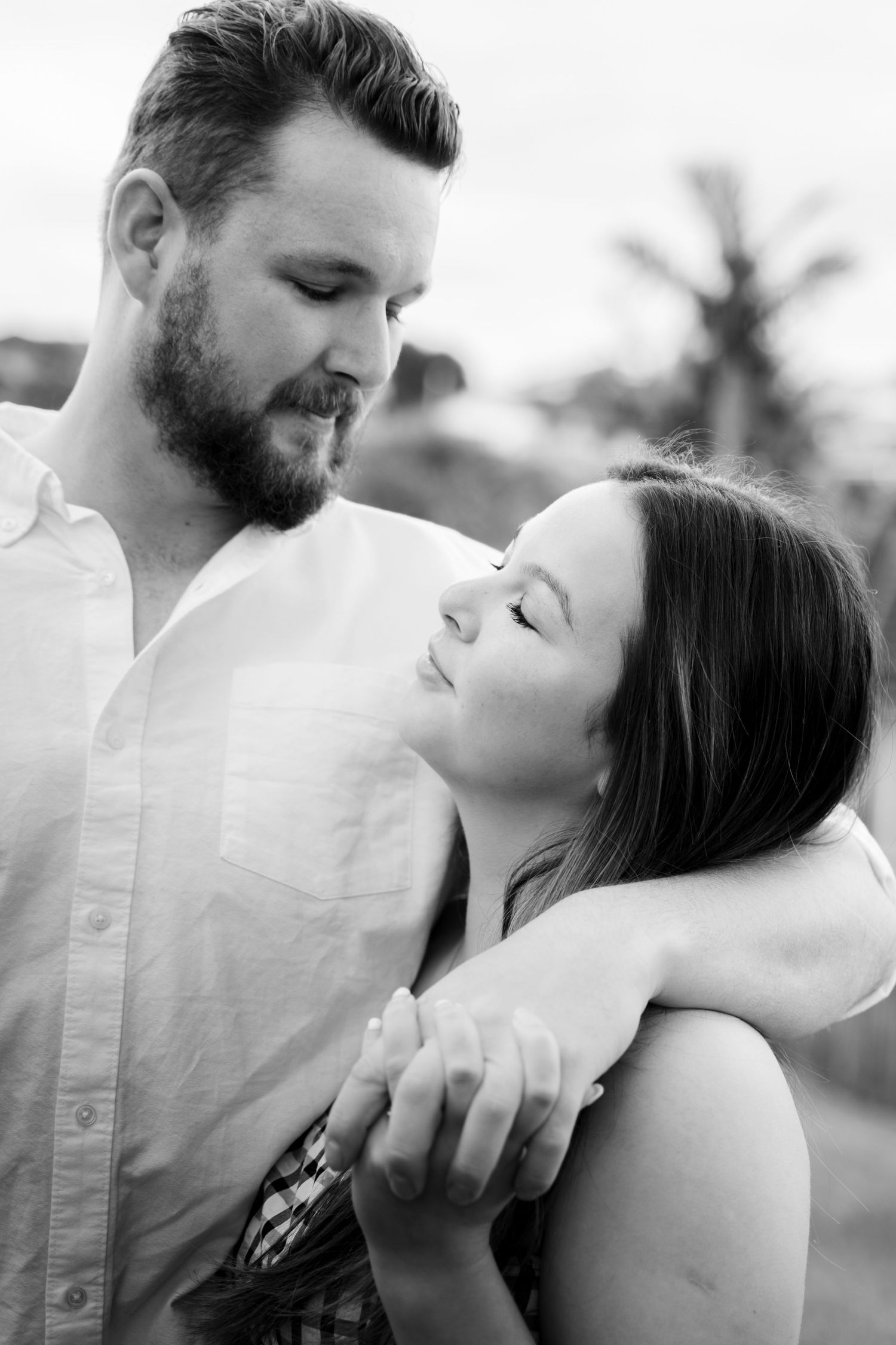 Black and white romantic couple portrait, man with beard embracing woman outdoors with palm trees in background.
