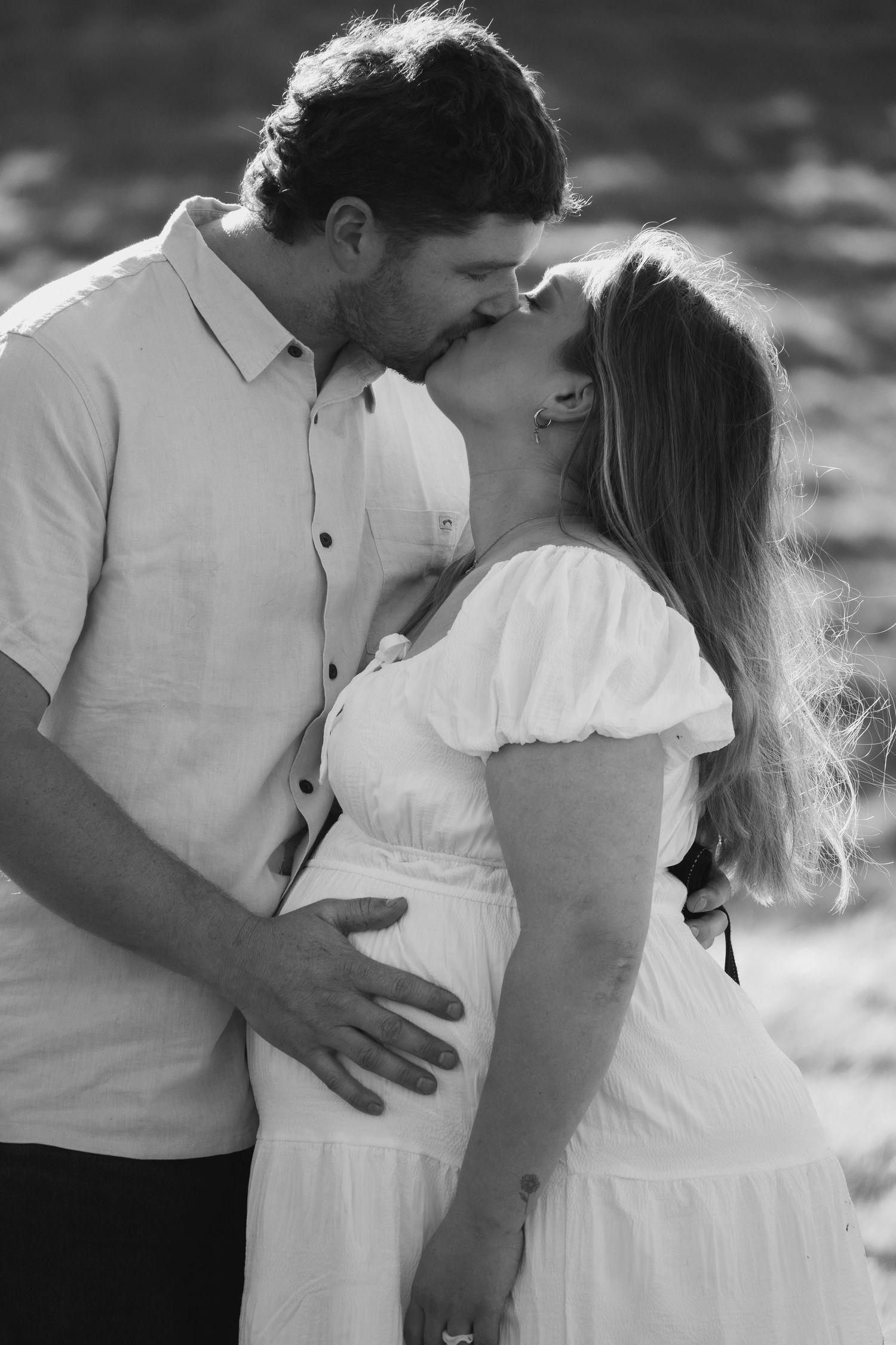 Black and white photo of a pregnant couple kissing outdoors, man tenderly holding woman's baby bump.