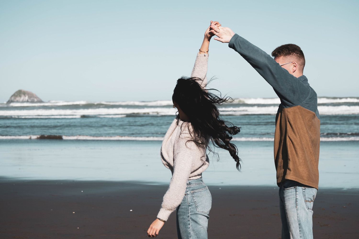 Couple dancing on a black sand beach by the ocean, man spinning woman with flowing dark hair.