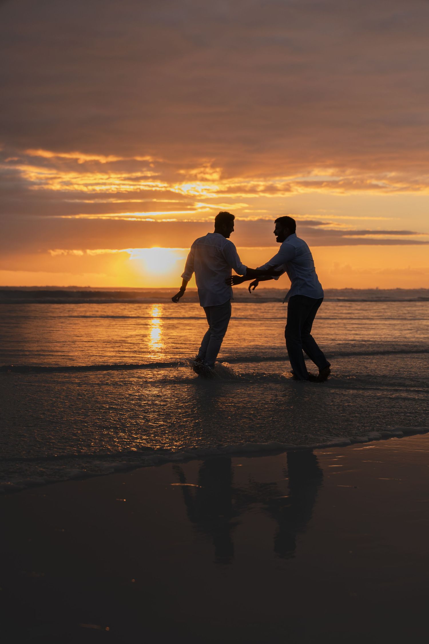 Two men holding hands and dancing on a beach at sunset, silhouetted against a vibrant orange sky.