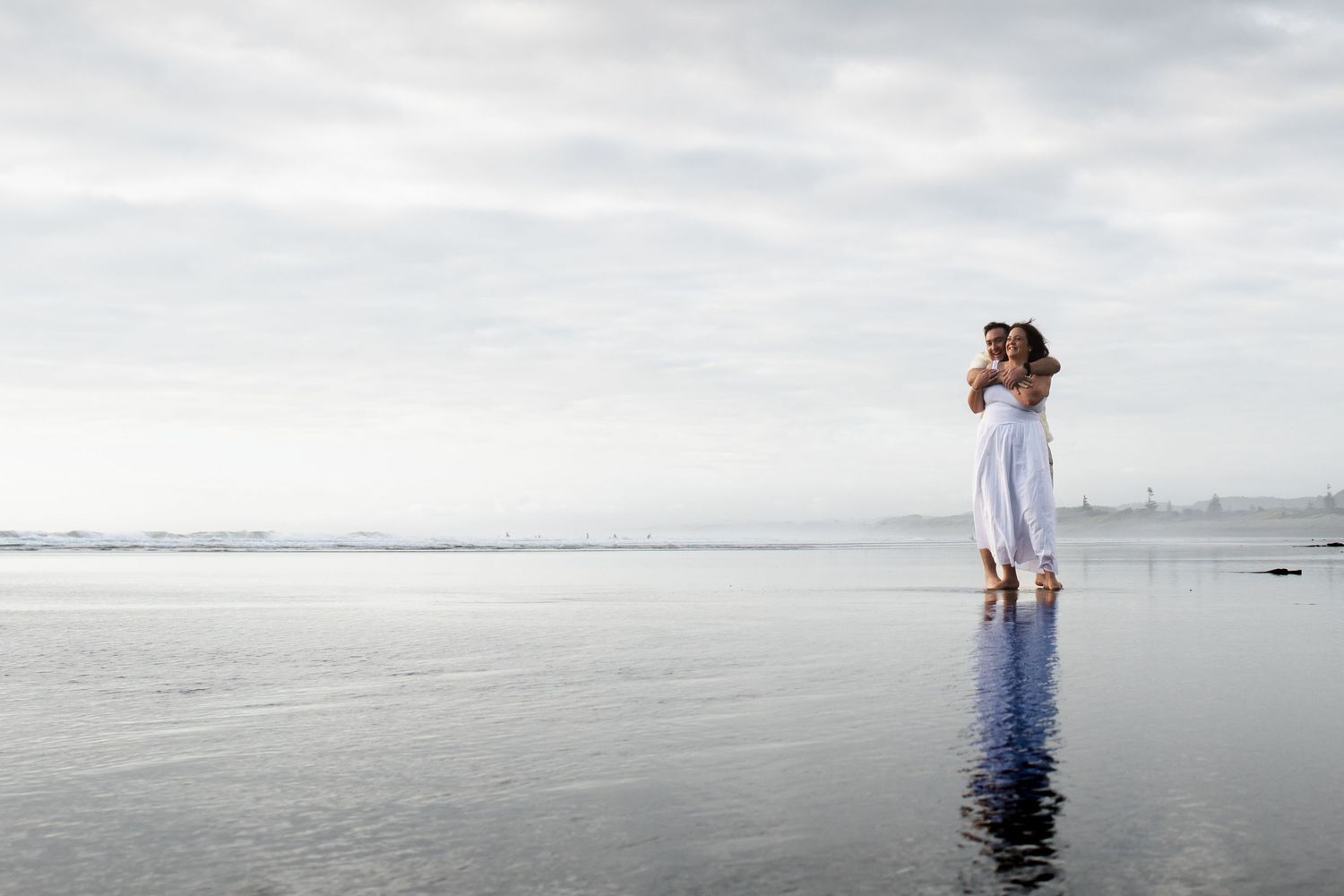 Couple embracing on a calm reflective beach with overcast skies, woman in white dress standing in shallow water.