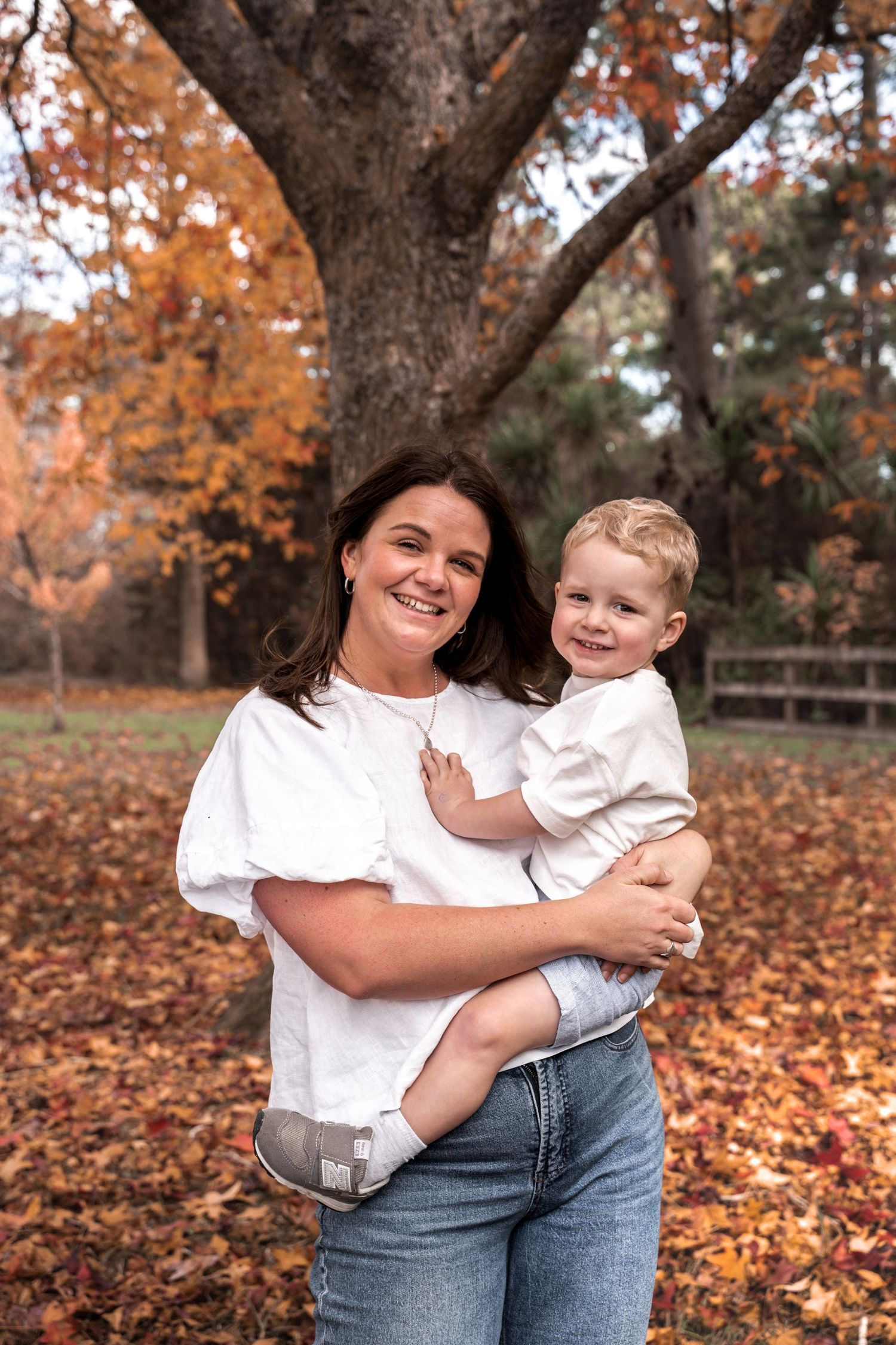 Smiling mother holding her young toddler son outdoors surrounded by colorful autumn fall foliage and trees.