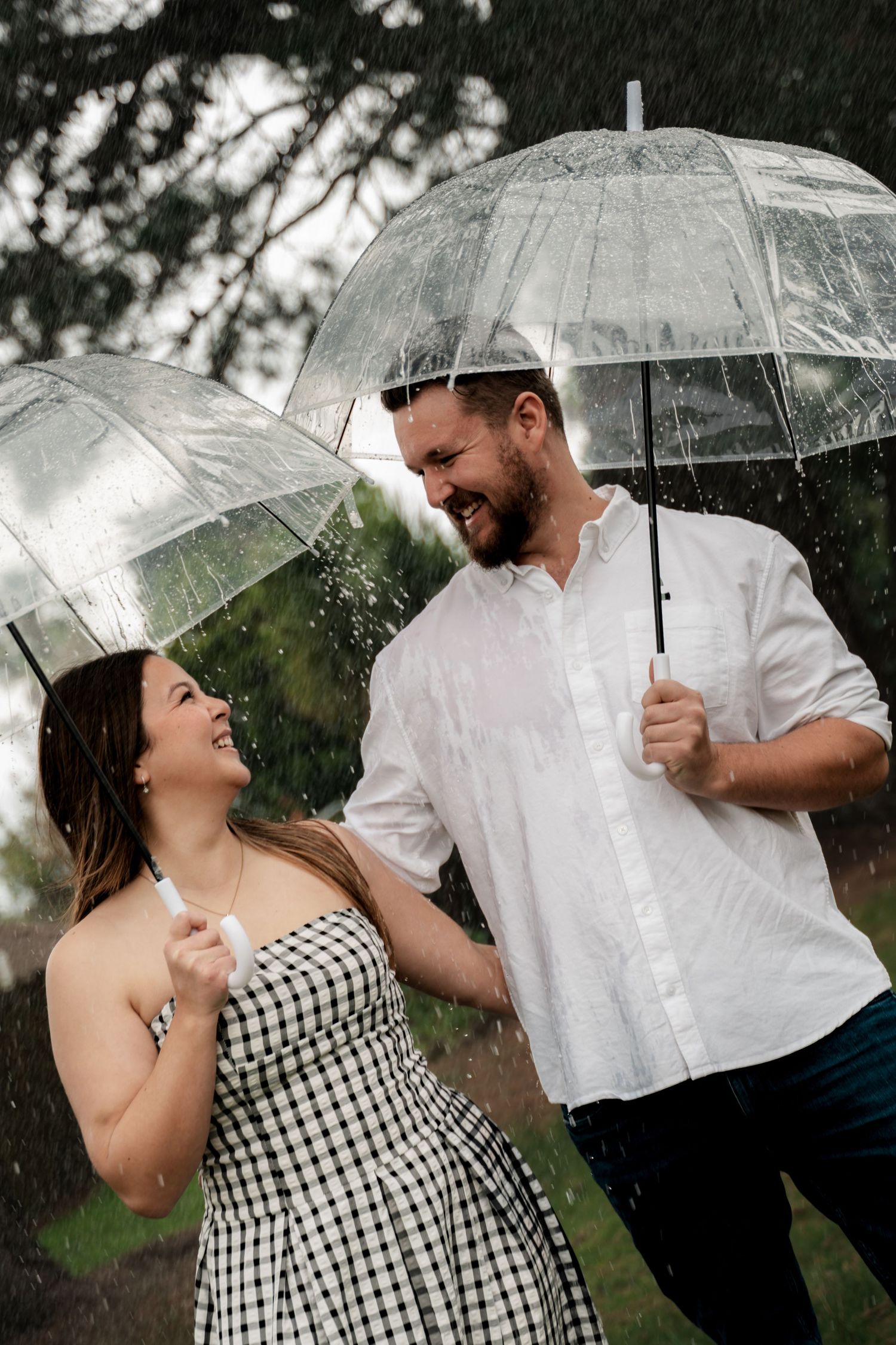 A laughing couple shares a romantic moment in the rain, each holding clear umbrellas, dressed in white and checkered outfits.