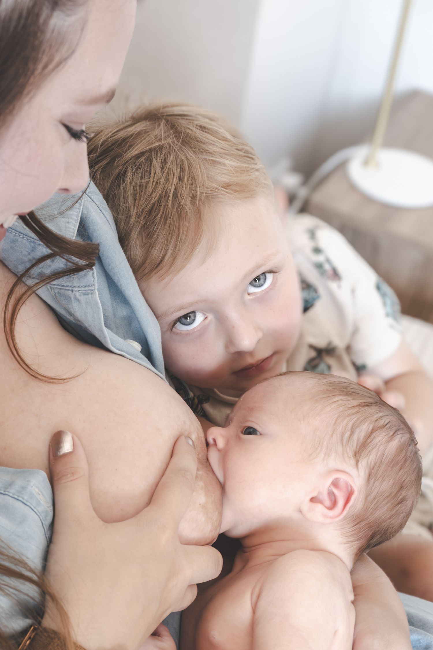 Mother breastfeeding newborn baby while toddler child watches closely in a warm, intimate home setting.