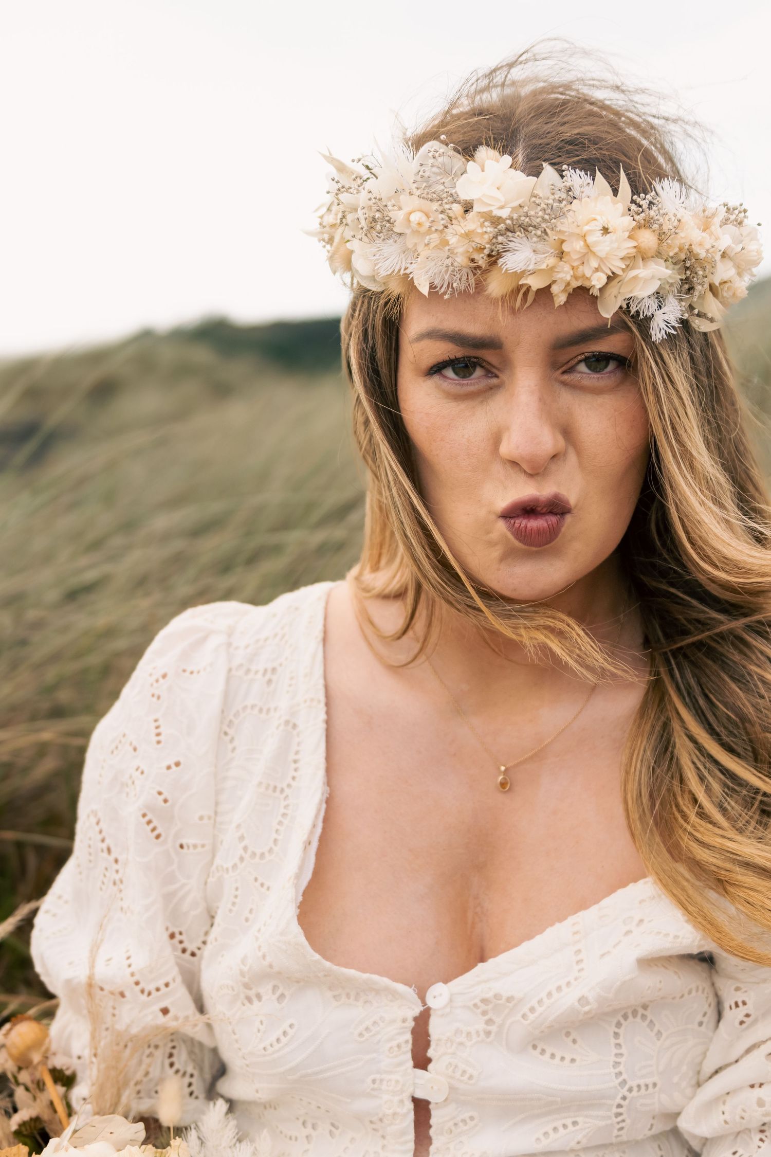 Young woman in white lace dress and floral crown posing outdoors with a playful expression.