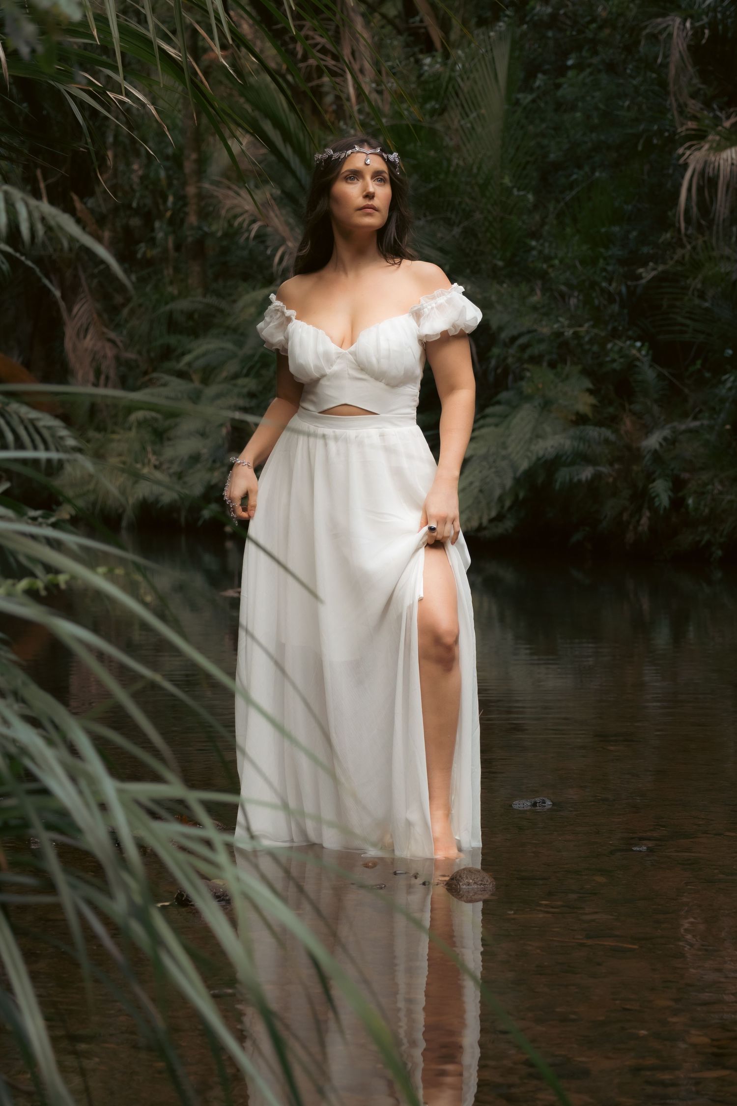 Woman in white off-shoulder bridal two-piece gown standing in a serene jungle river surrounded by lush greenery.