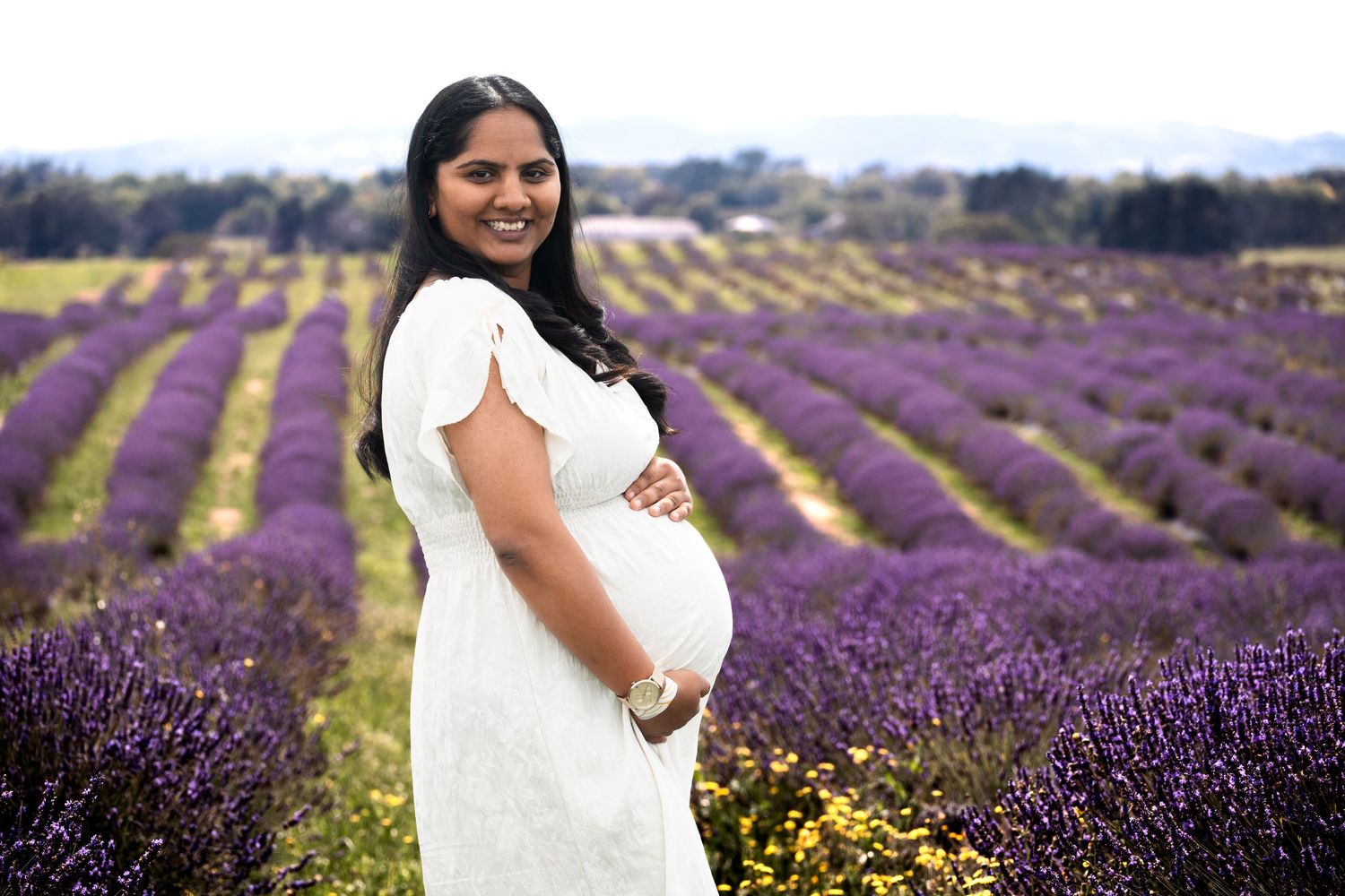 Pregnant woman in white dress smiling in a lavender field during a maternity photoshoot.
