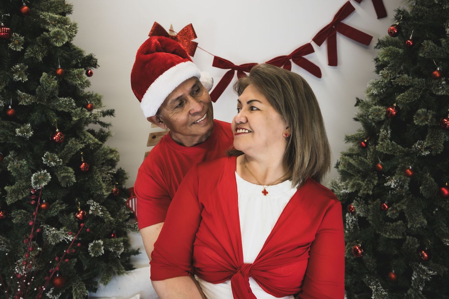 A couple in red Christmas outfits smiling together between decorated Christmas trees, man wearing a Santa hat.