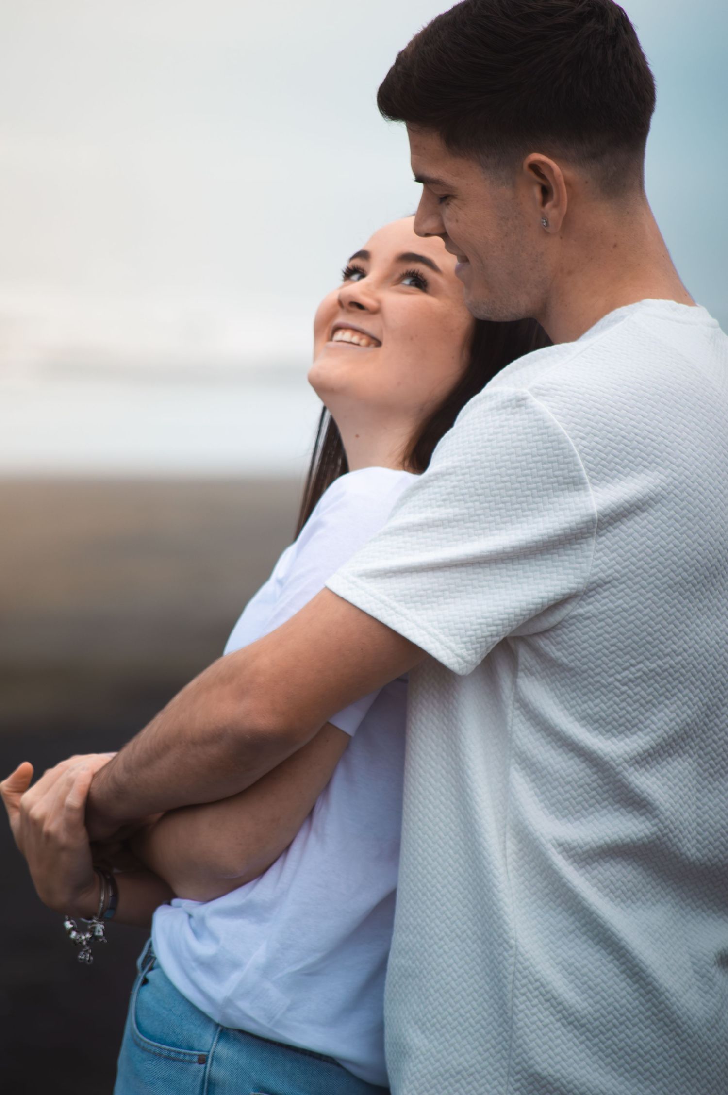 Romantic couple embracing outdoors, woman smiling up at man in white t-shirts near ocean backdrop.