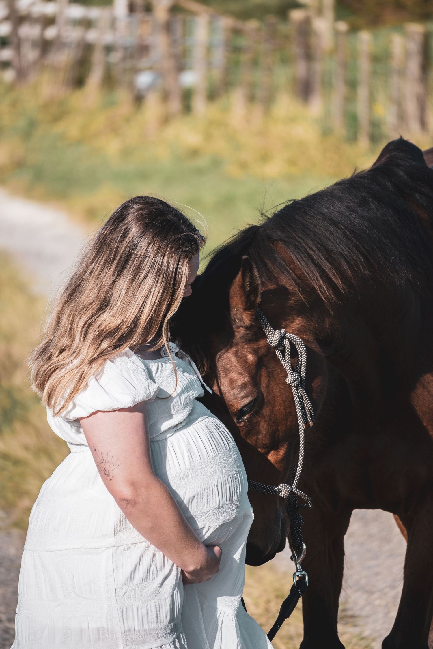 Pregnant woman in white dress tenderly bonding with a dark brown horse on a sunny rural path.