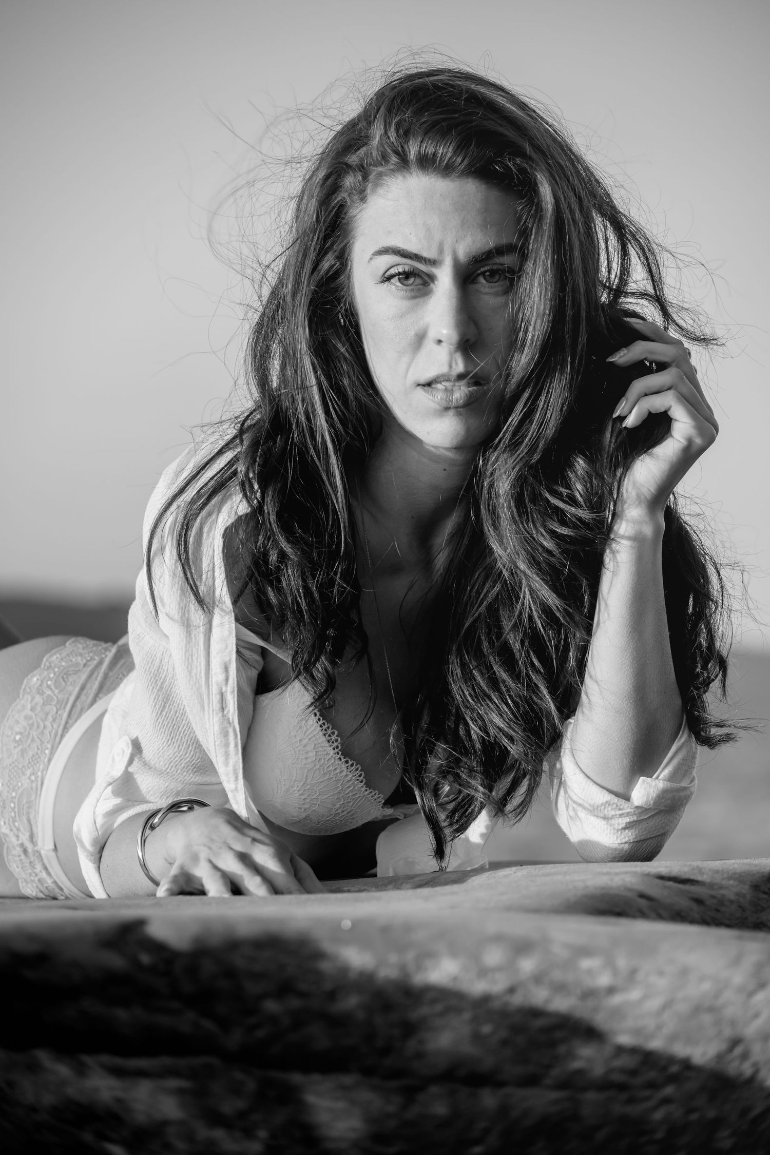 Black and white boudoir photo of a woman with long wavy hair posing on a bed in an open white shirt.