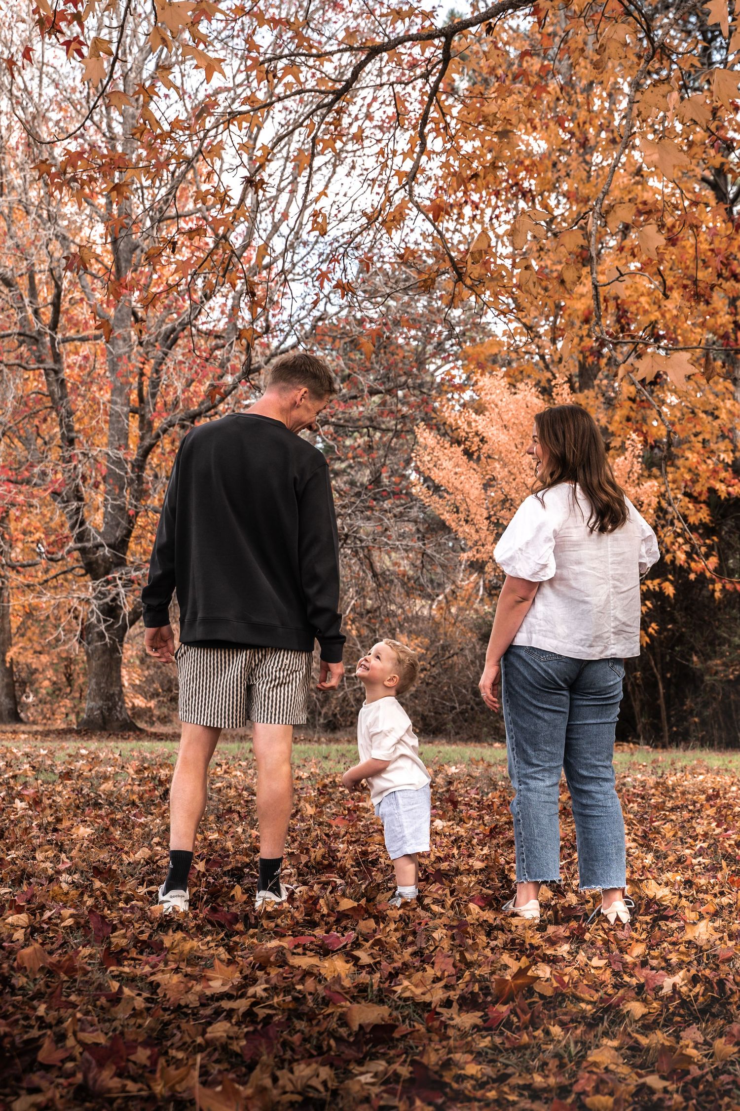 Family of three enjoying autumn park, parents and toddler standing among fallen leaves with colorful fall trees.