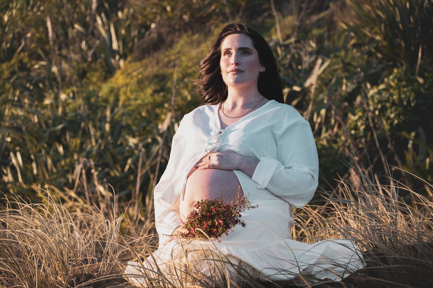 Pregnant woman in white dress sitting in golden grass field, cradling her baby bump with a serene expression.