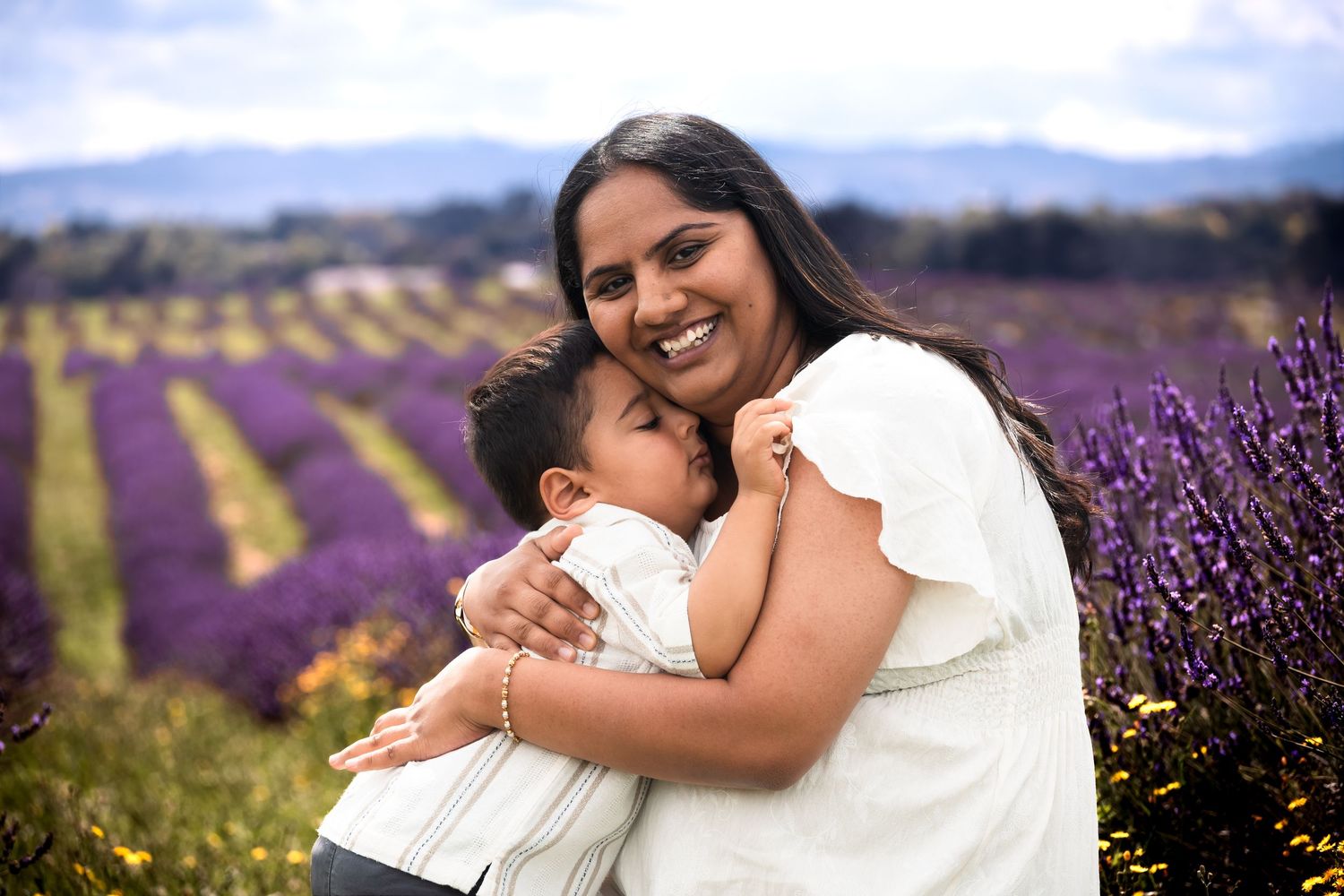 A smiling woman hugs a young child in a vibrant purple lavender field with mountains in the background.