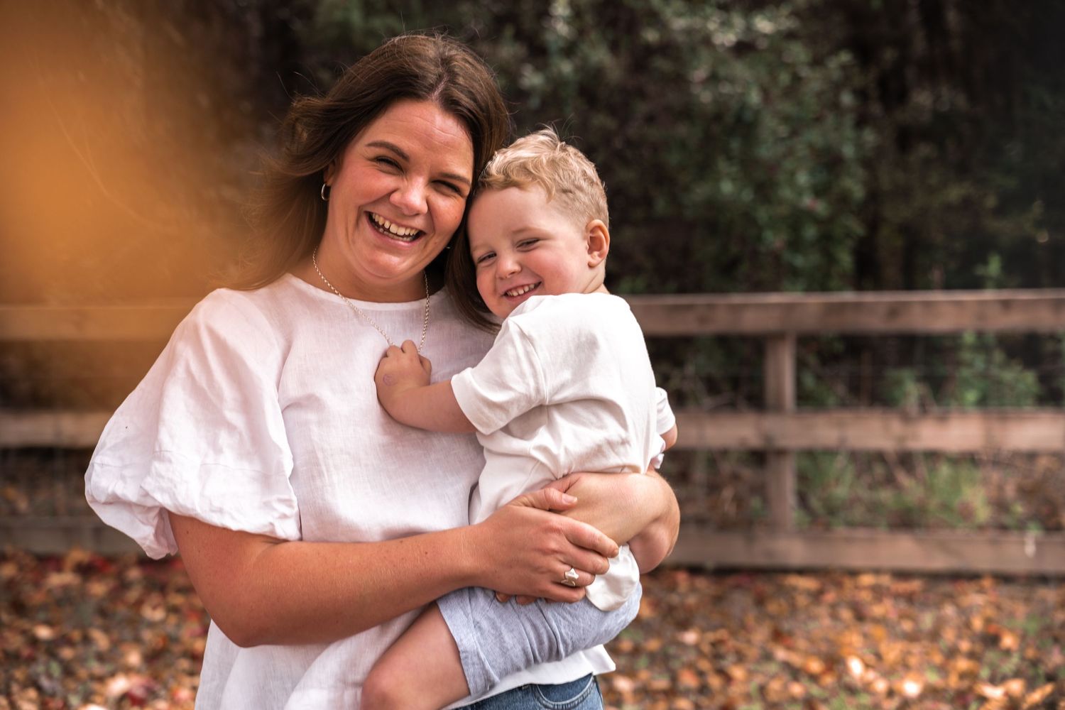 A smiling woman in a white top holds a happy toddler boy outdoors in a wooded park setting.
