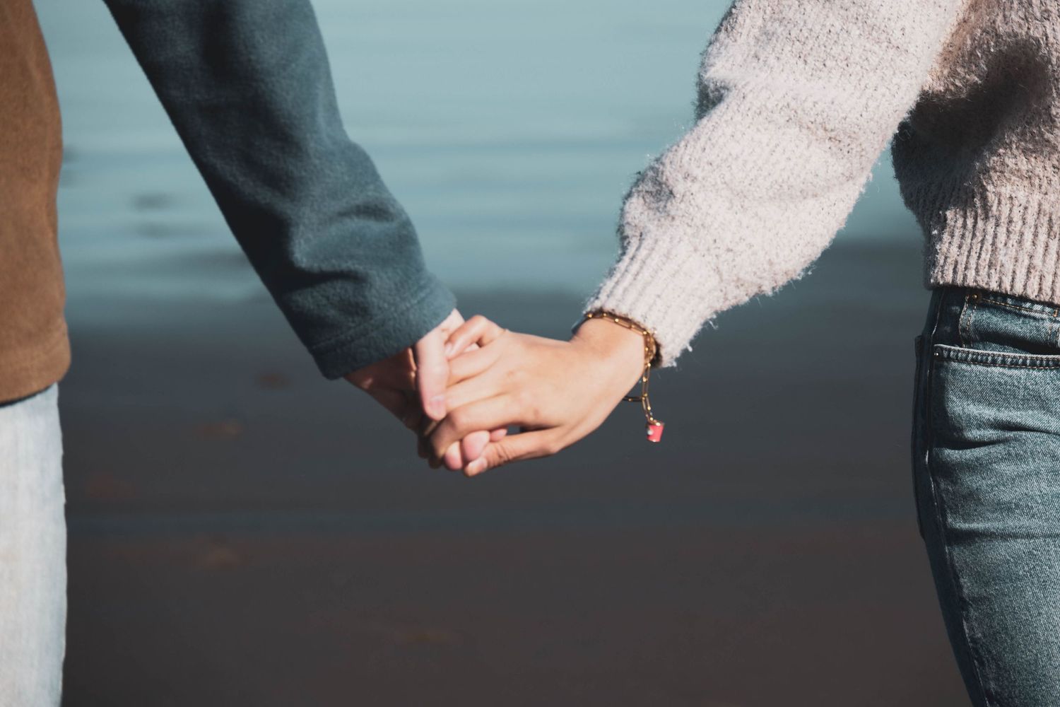 Couple holding hands near water, wearing casual sweaters and jeans, showcasing a romantic connection outdoors.