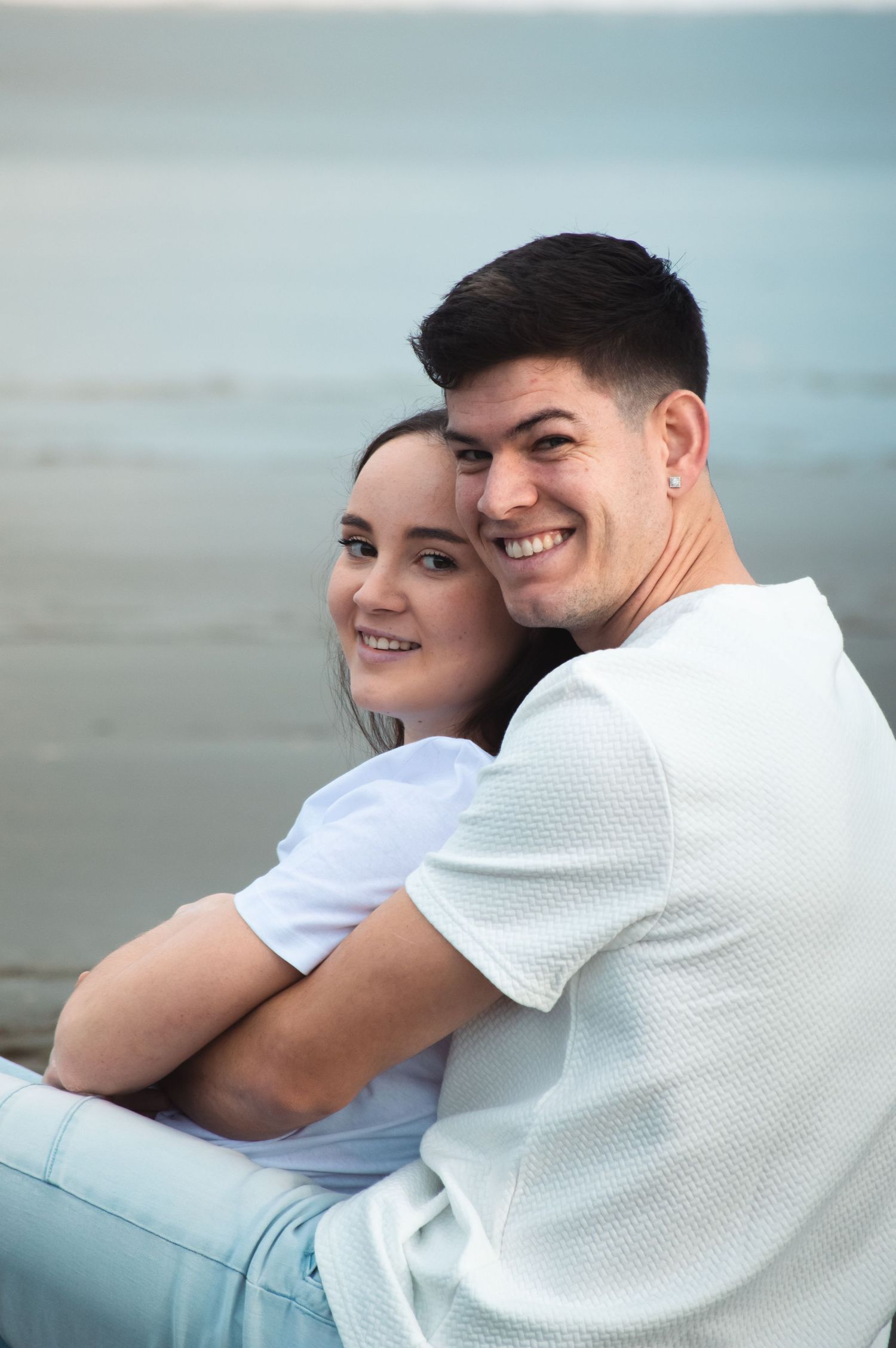 A smiling couple sitting together on a beach, the man embracing the woman from behind, both wearing white tops.