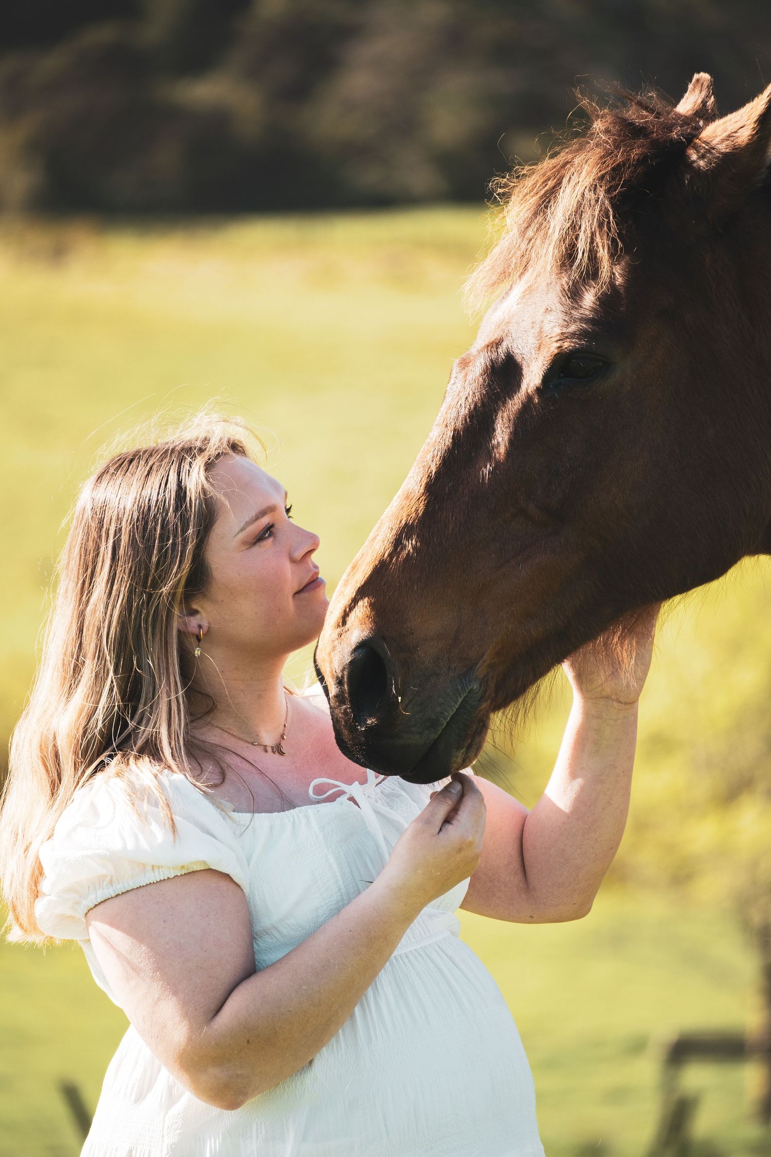 Pregnant woman in white dress tenderly touching a chestnut horse's nose in a sunlit green field.