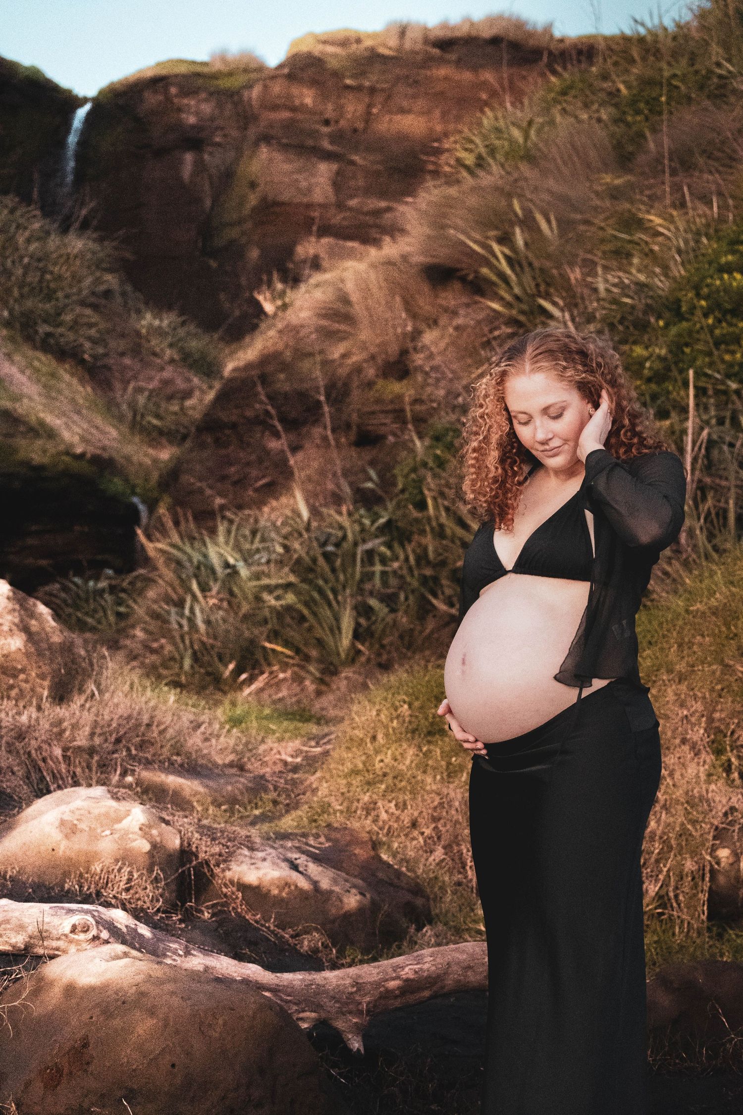 Pregnant woman in black dress cradling her baby bump outdoors near a waterfall and rocky cliff.