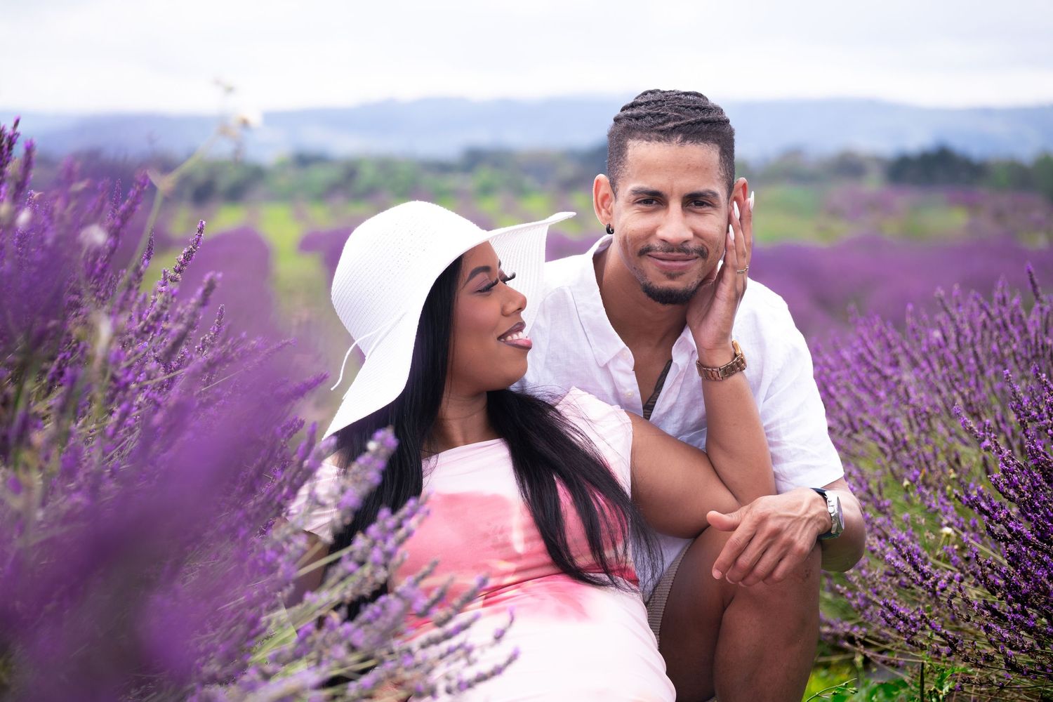 Couple posing romantically in a vibrant purple lavender field, woman in white hat, man in white shirt.