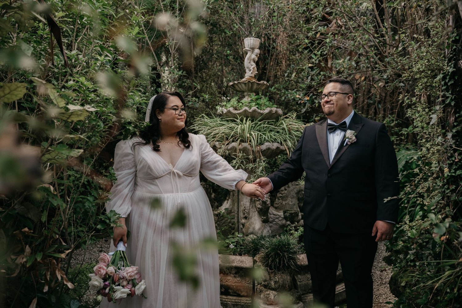 Bride and Groom holding hands in a lush garden, bride in white gown and groom in black tuxedo, at a west auckland wedding venue.