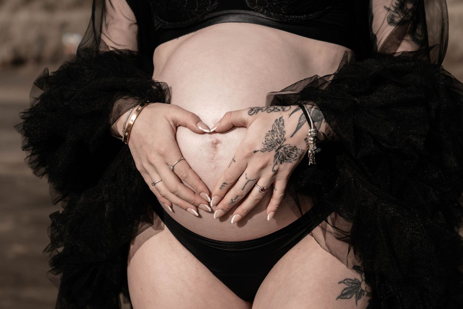 Pregnant woman in black lingerie forming heart shape on baby bump with tattooed hands.