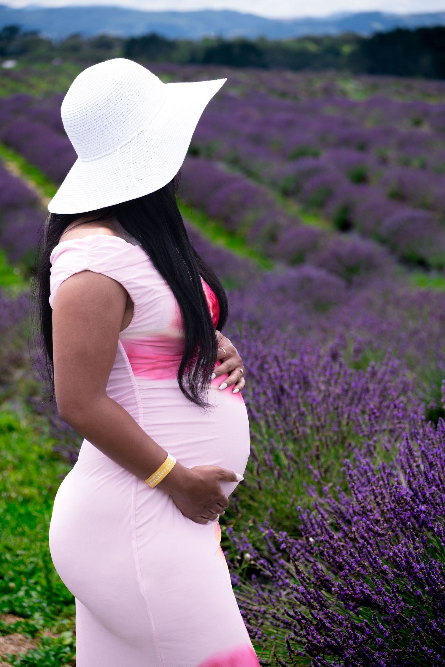Pregnant woman in pink dress and white sun hat cradling baby bump in a blooming purple lavender field.