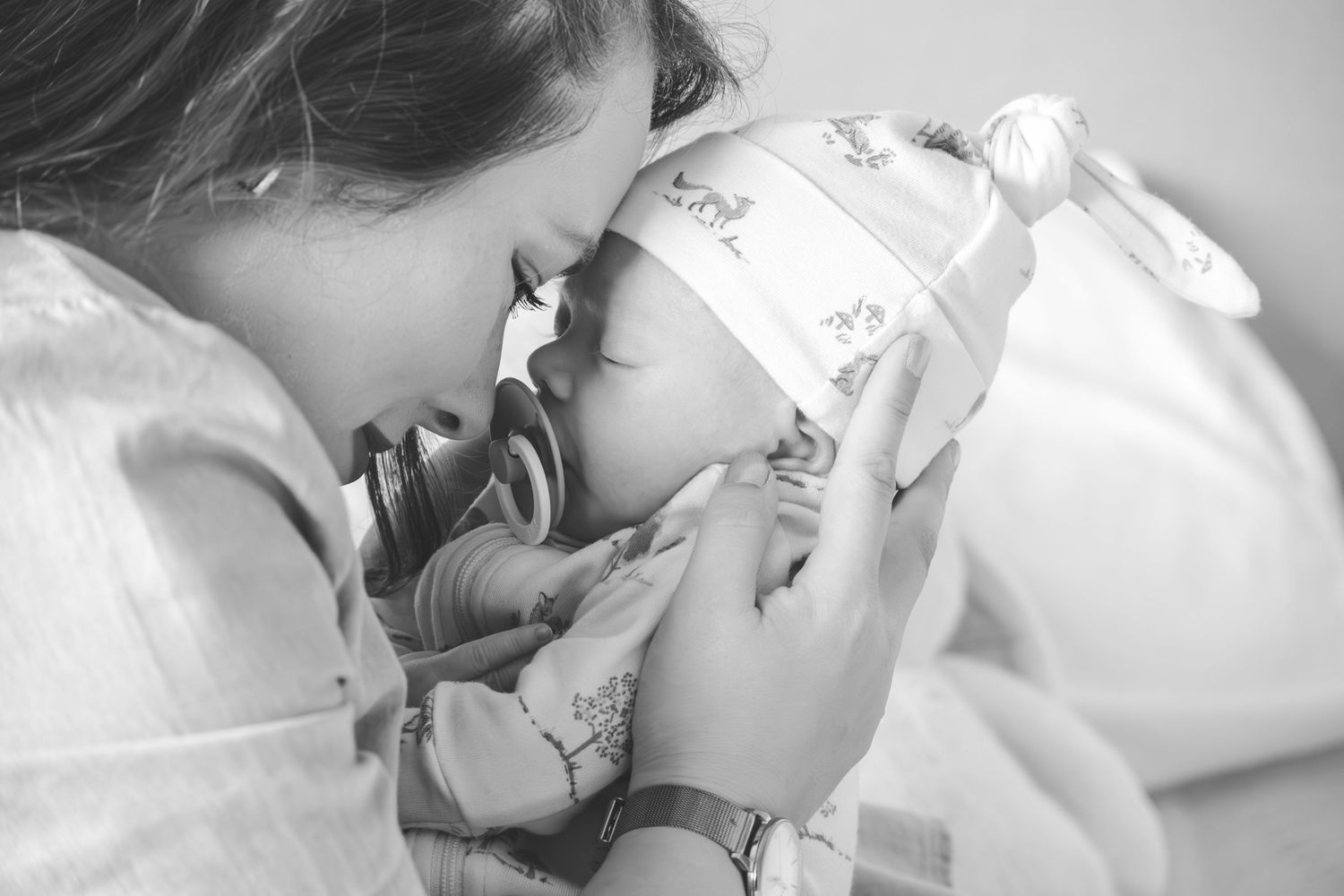 Black and white photo of a mother tenderly kissing her newborn baby wrapped in a floral hat.