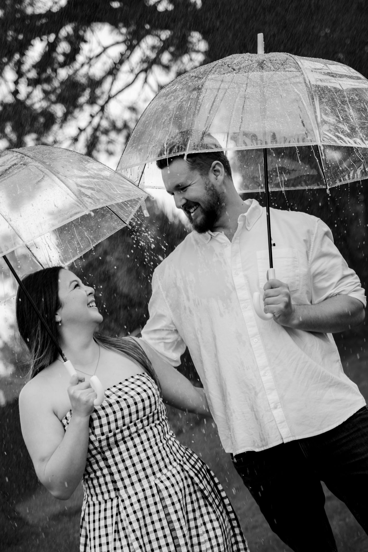 Black and white photo of a laughing couple holding clear umbrellas together in the rain.