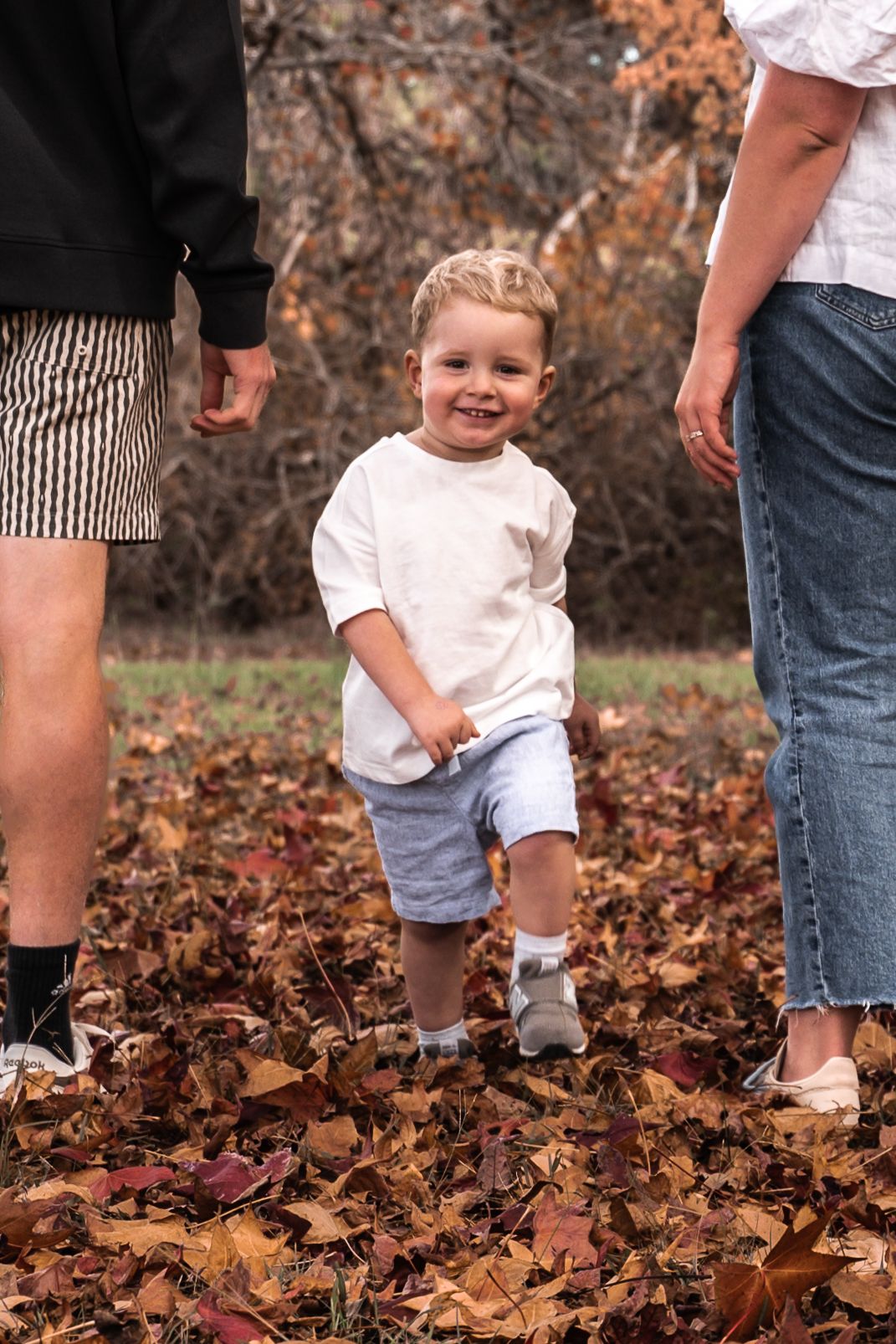 A happy toddler boy plays in autumn leaves between two adults during a fall family outdoor photo session.