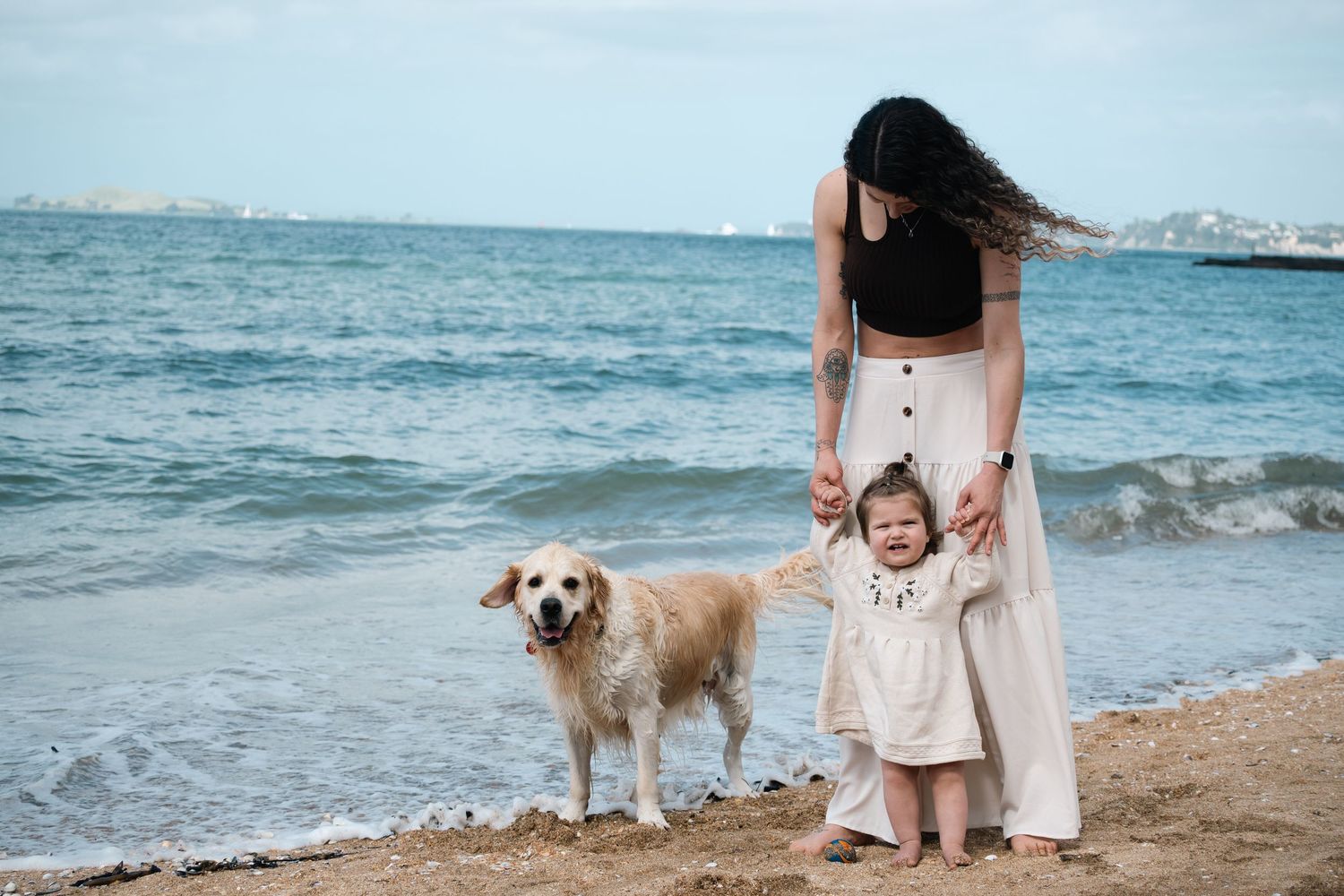 Woman and toddler holding hands on sandy beach with golden retriever by ocean waves.